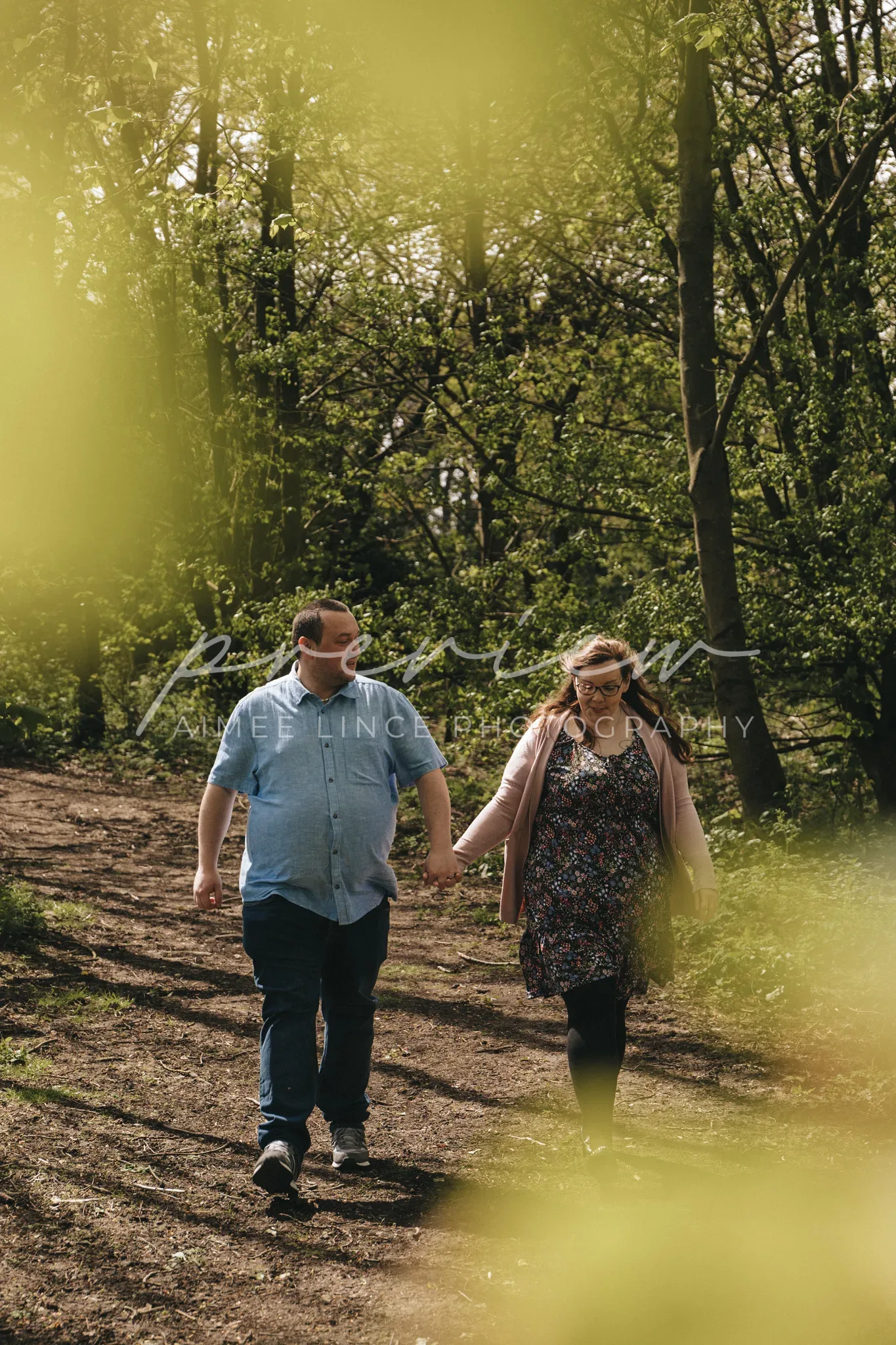A couple holding hands and walking on a sunlit forest path. Ashley, wearing a floral dress, and the man, in a blue shirt and jeans, smile as they walk. Sunlight filters through