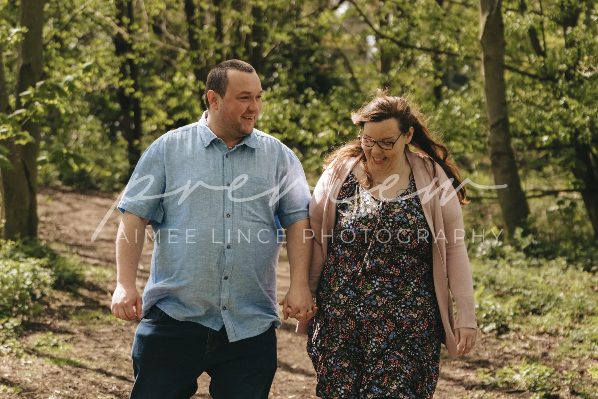 A couple holding hands and smiling while walking through a sunlit forested area, with the man on the left wearing a blue shirt and Ashley wearing a floral dress and glasses.