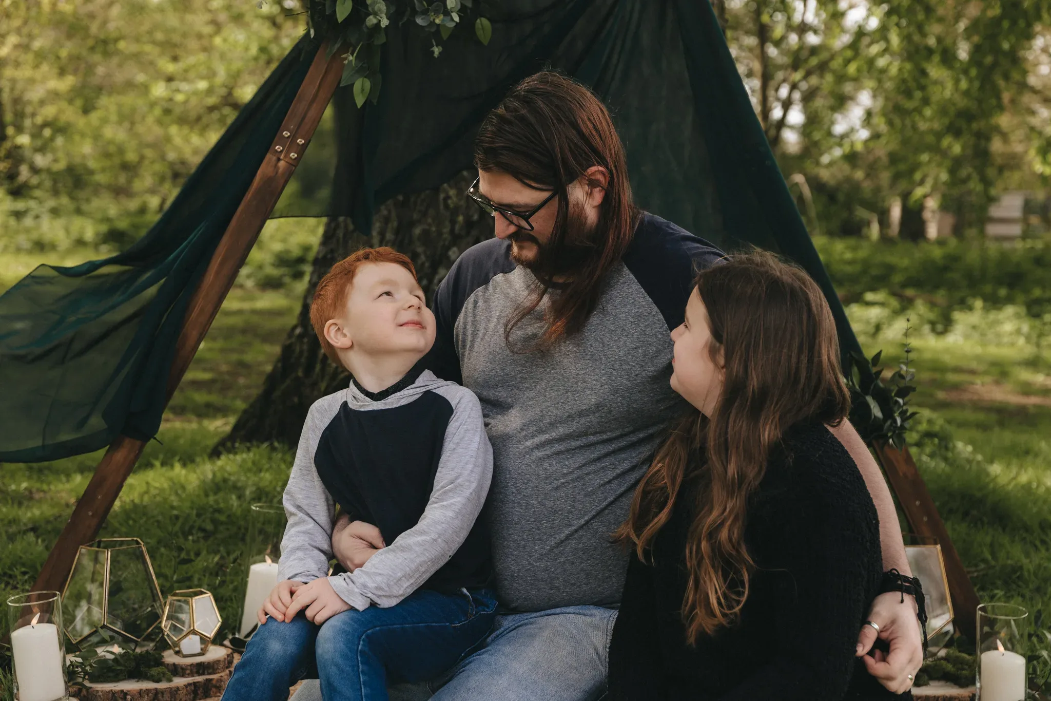 A father with long hair and a beard sits with his son and daughter on a picnic blanket under a tent in a park, sharing a joyful moment. the area is decorated with candles and greenery.