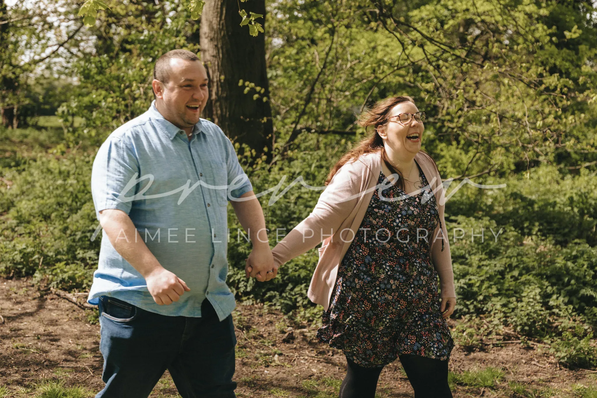 A joyful man and woman holding hands and running through a sunlit park. Both are laughing and dressed in casual spring attire. Trees with fresh green leaves and grassy areas are visible in the background.