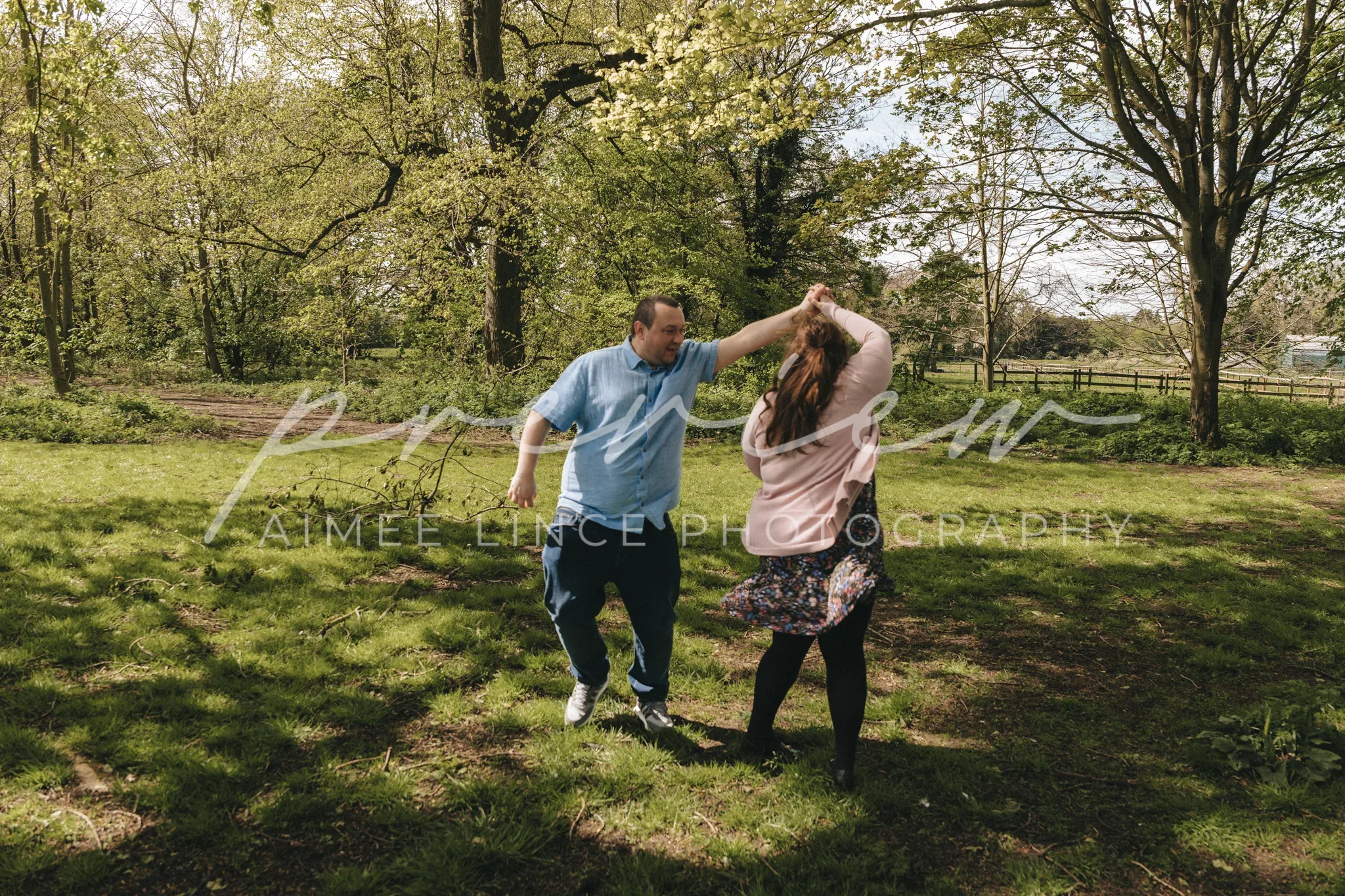 A man and a woman dance joyfully in a lush park under a canopy of trees, with sunlight filtering through the leaves. Samantha flicks her hair, and both are smiling, captured mid-twirl