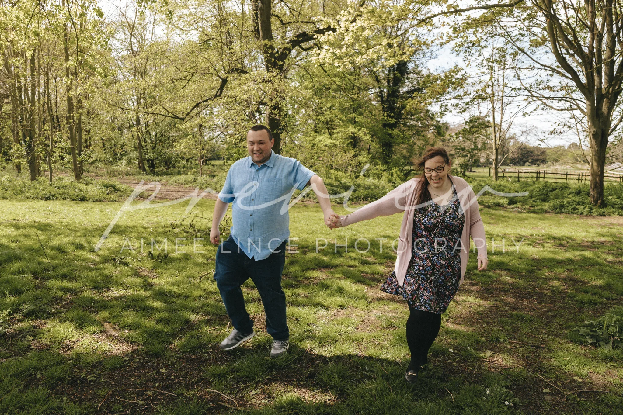 A joyful couple holding hands and running through a lush, green park, with blossoming trees and vibrant grass under a clear sky. Samantha is dressed in a blue shirt and jeans, while Ashley wears a
