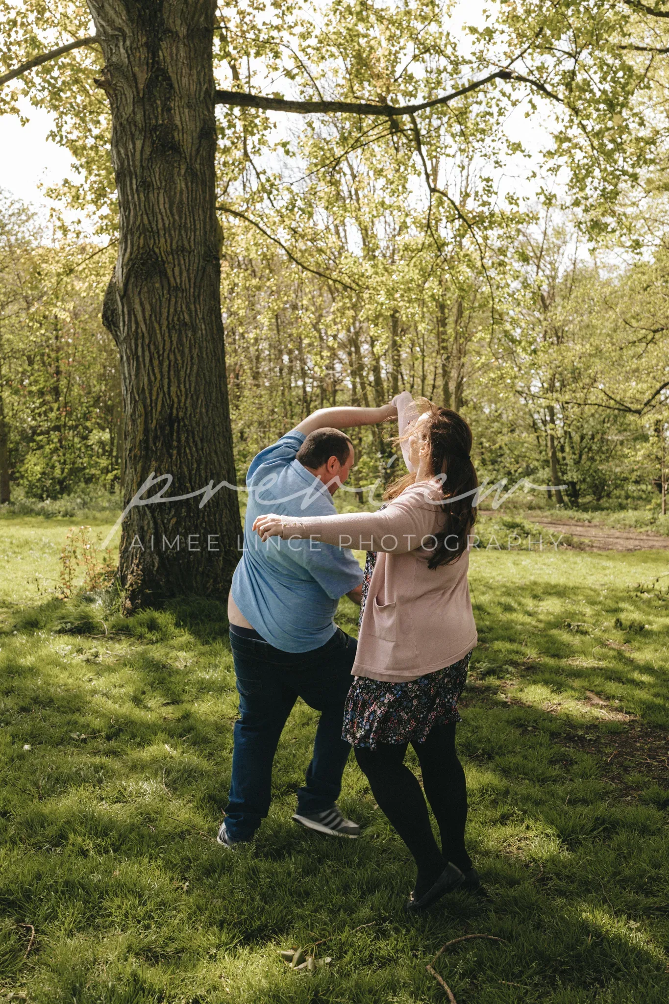 A man and Samantha playfully dance under a large tree in a sunny park. Samantha swings her long hair as the man reaches out to her. They both wear casual spring clothing and are surrounded by lush