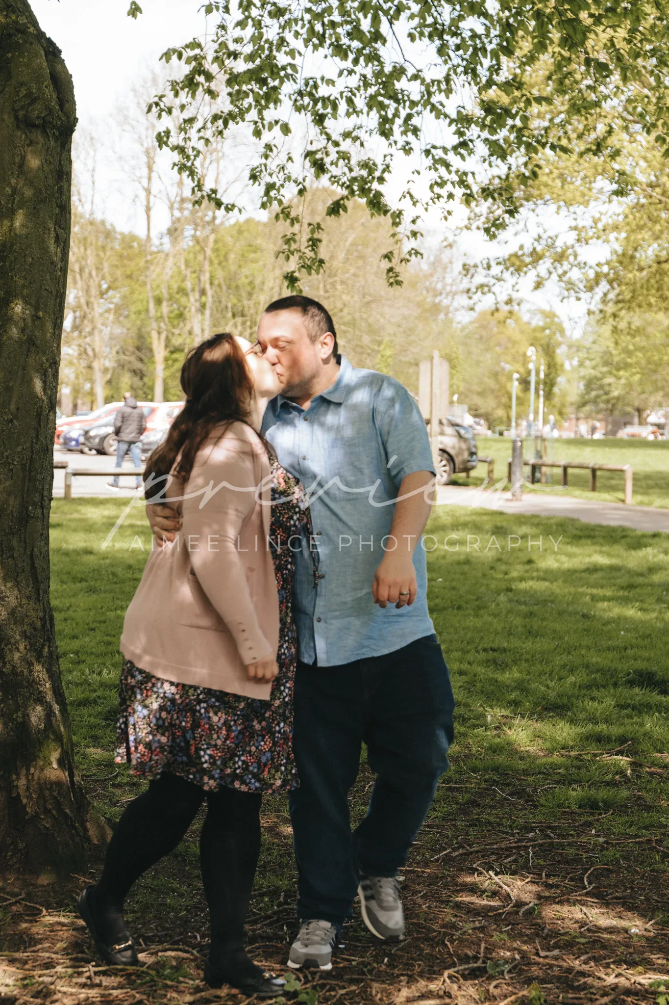 A couple shares a kiss under a tree in a sunny park setting during their wedding photography session. The woman, wearing a floral dress and black leggings, embraces a man in a blue shirt and jeans.