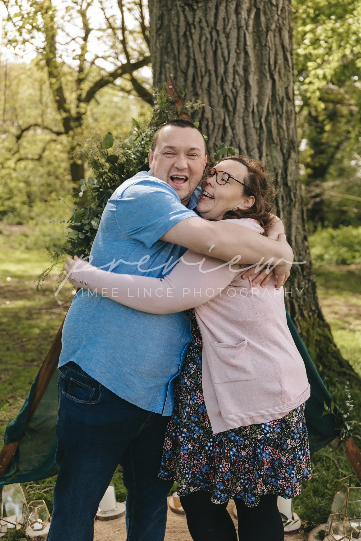 A joyful couple embraces under a tree in a lush park. Ashley, wearing a blue shirt and jeans, and Samantha, sporting a floral dress and glasses, are smiling broadly, radiating happiness.