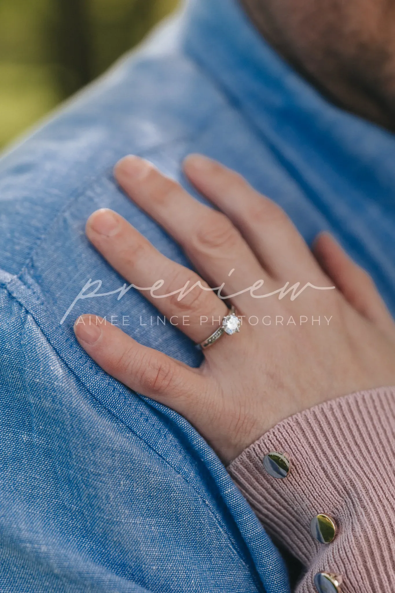 A woman's hand adorned with an engagement ring rests on a man's shoulder, who is wearing a blue shirt. The focus is on the sparkling ring against the blurred background of greenery. The image