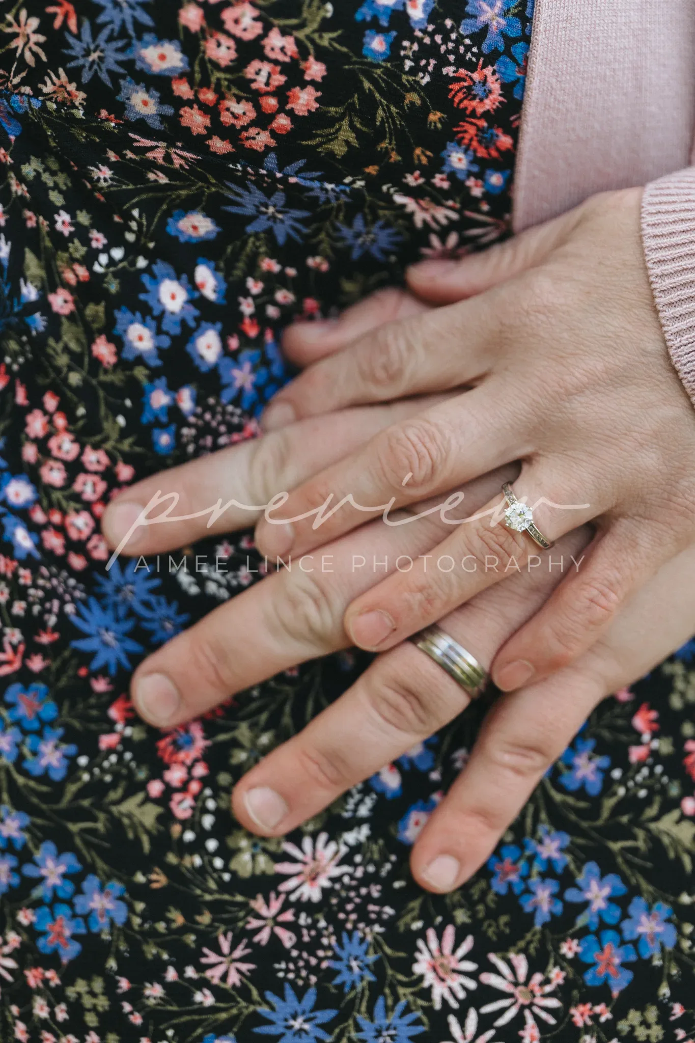 A close-up image of a couple's hands on a floral background. The woman's hand has a sparkling engagement ring, placed atop the man's hand that features a wedding band. The watermark reads