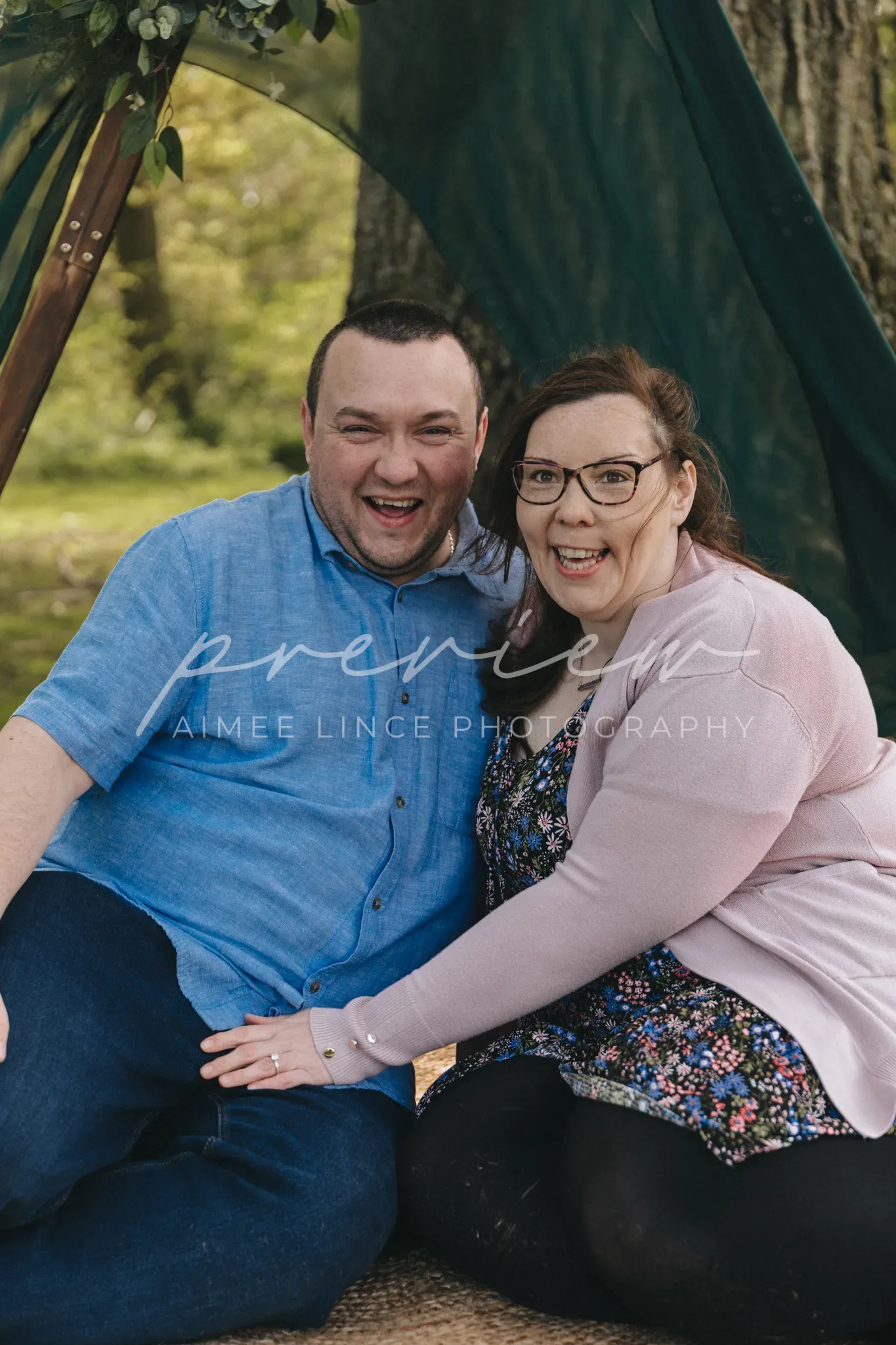A happy couple sits closely under a tree with a decorative green fabric draped above. The man, in a blue shirt, and the woman, named Samantha, in a cardigan and floral dress, are