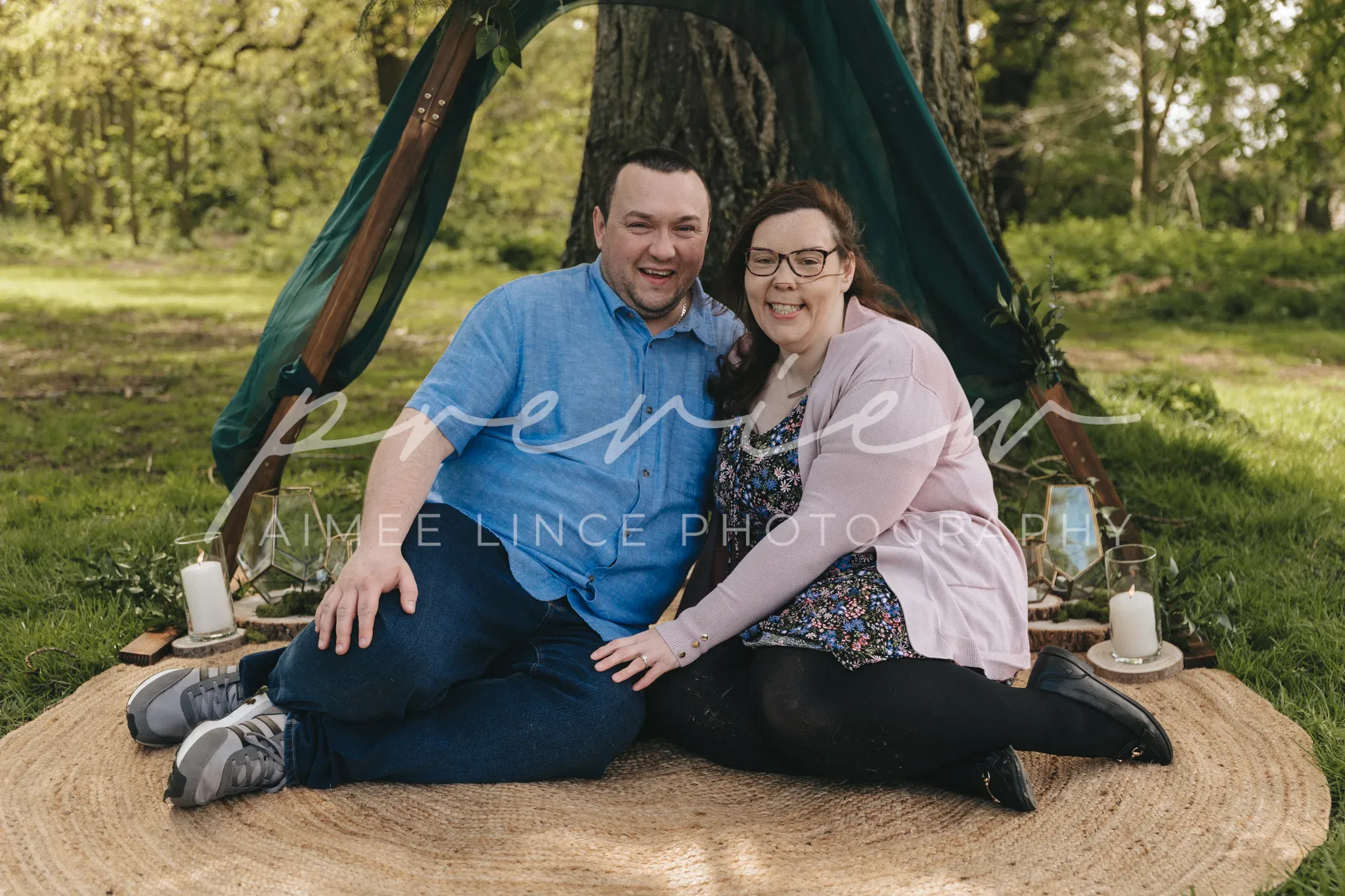 A smiling couple, Ashley and Samantha, sit on a circular rug under a tent-like structure in a lush park. Ashley wears a blue shirt and jeans; Samantha wears a floral dress and cardigan.