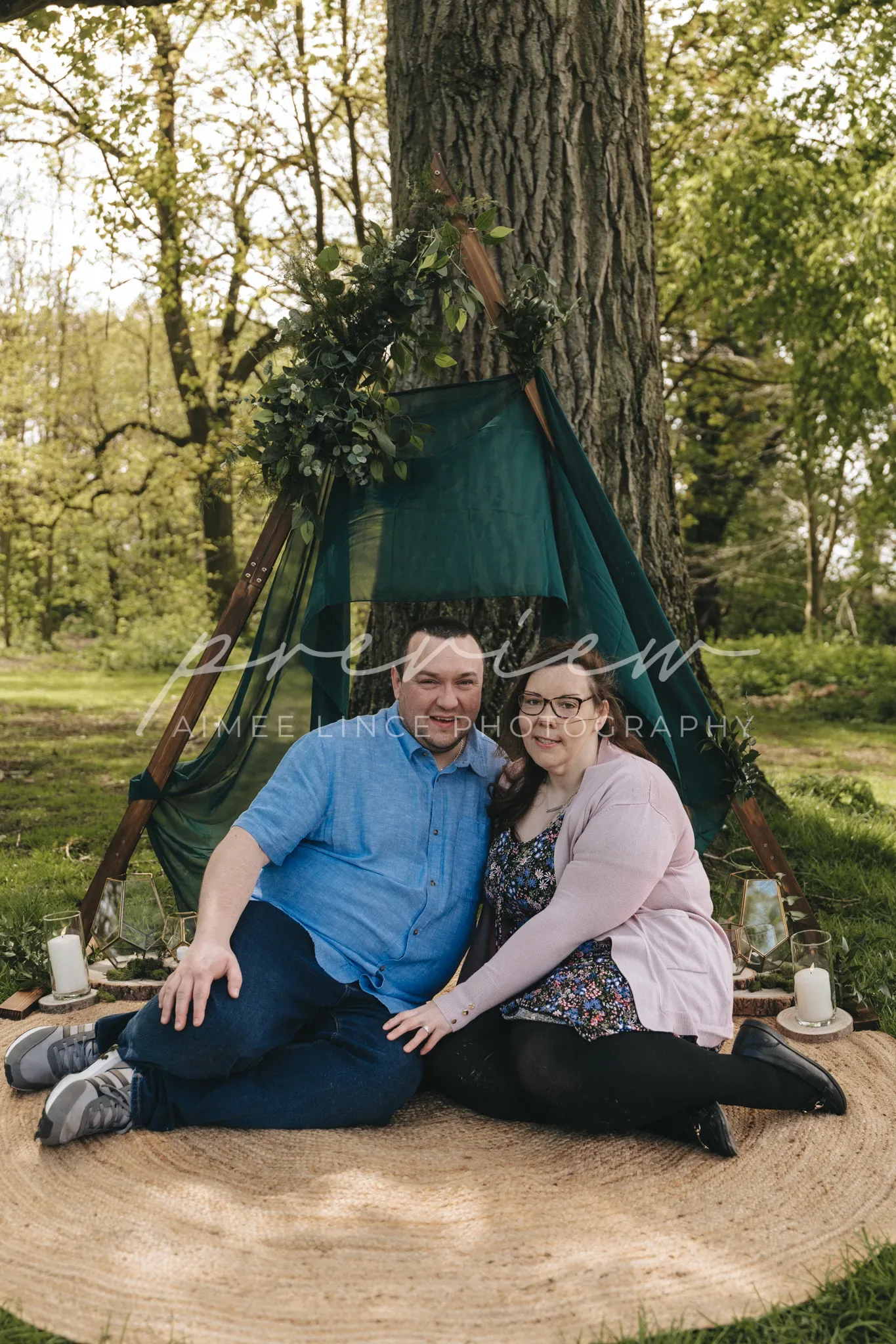 A couple, Ashley and Samantha, sit closely on a circular mat under a teepee adorned with greenery in a lush park, smiling at the camera. Lanterns and natural decor enhance the cozy,