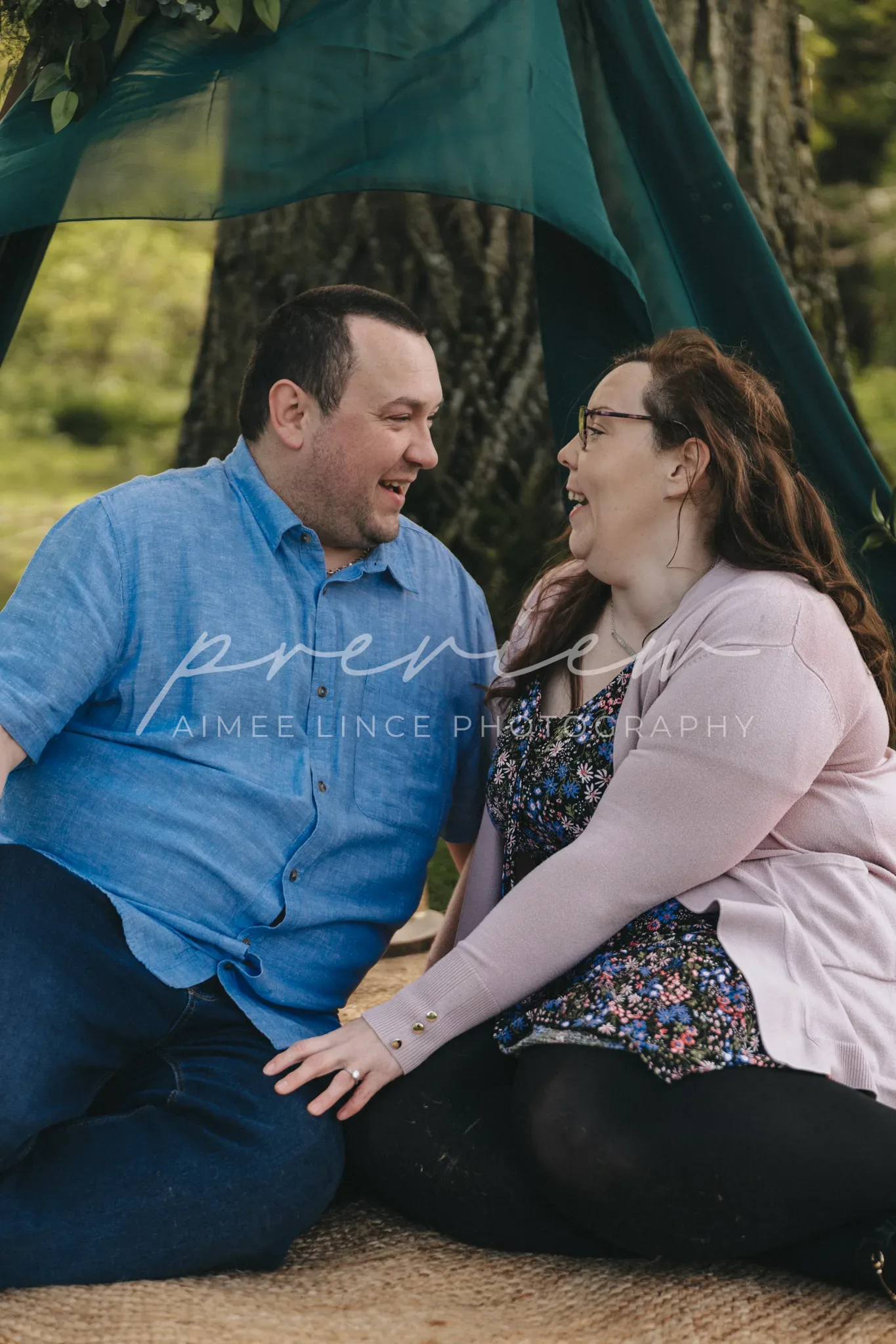A man and a woman, Ashley and Samantha, sit on a blanket, smiling joyfully at each other under a forest canopy. Both wear casual outfits; the man in a blue shirt and Ashley in