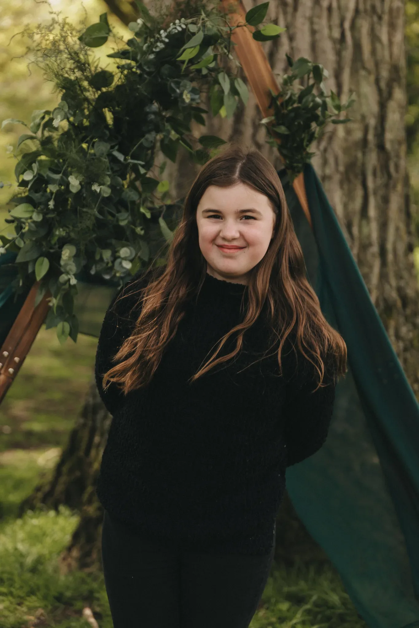 A young girl with long brown hair and wearing a black sweater smiles at the camera, standing in front of a green tent and a decorative greenery arch in a lush, outdoor setting.
