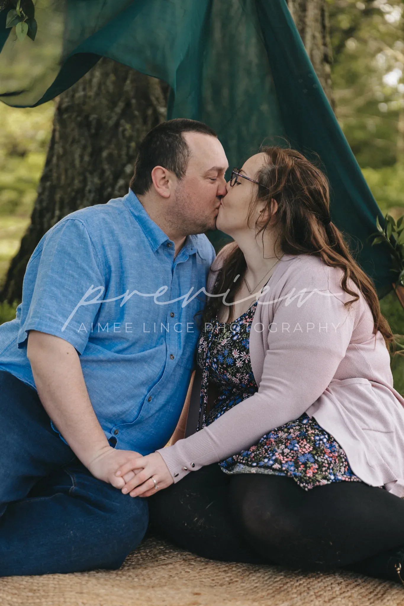 A couple sits on a blanket under a tree, kissing. The man wears a blue shirt, and the woman, named Ashley, wears a pink cardigan over a floral dress. The environment suggests a