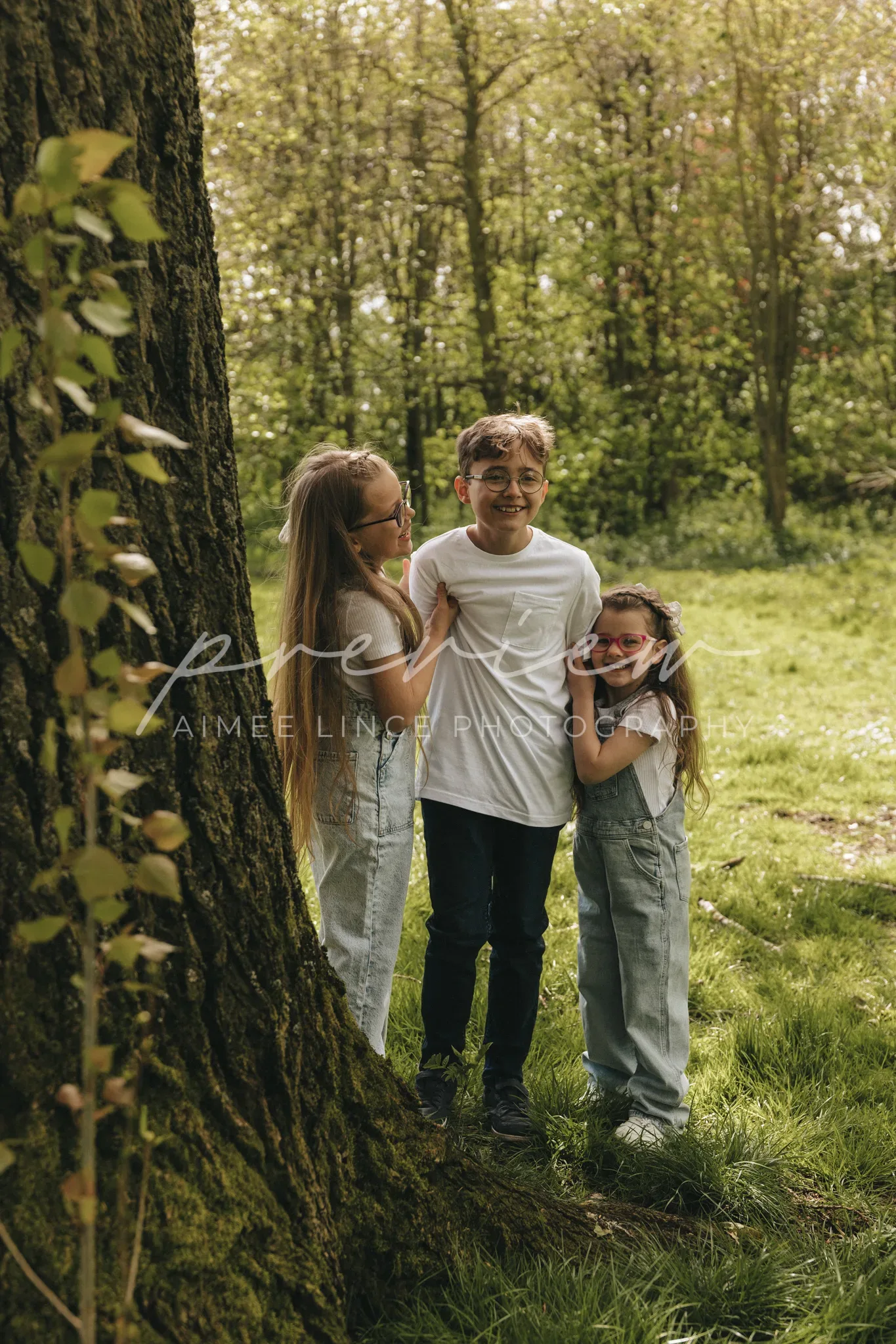 Three children laughing together in a lush green forest. two girls lean on a tree on either side of a boy, all in casual attire, surrounded by vibrant greenery and soft sunlight filtering through leaves.