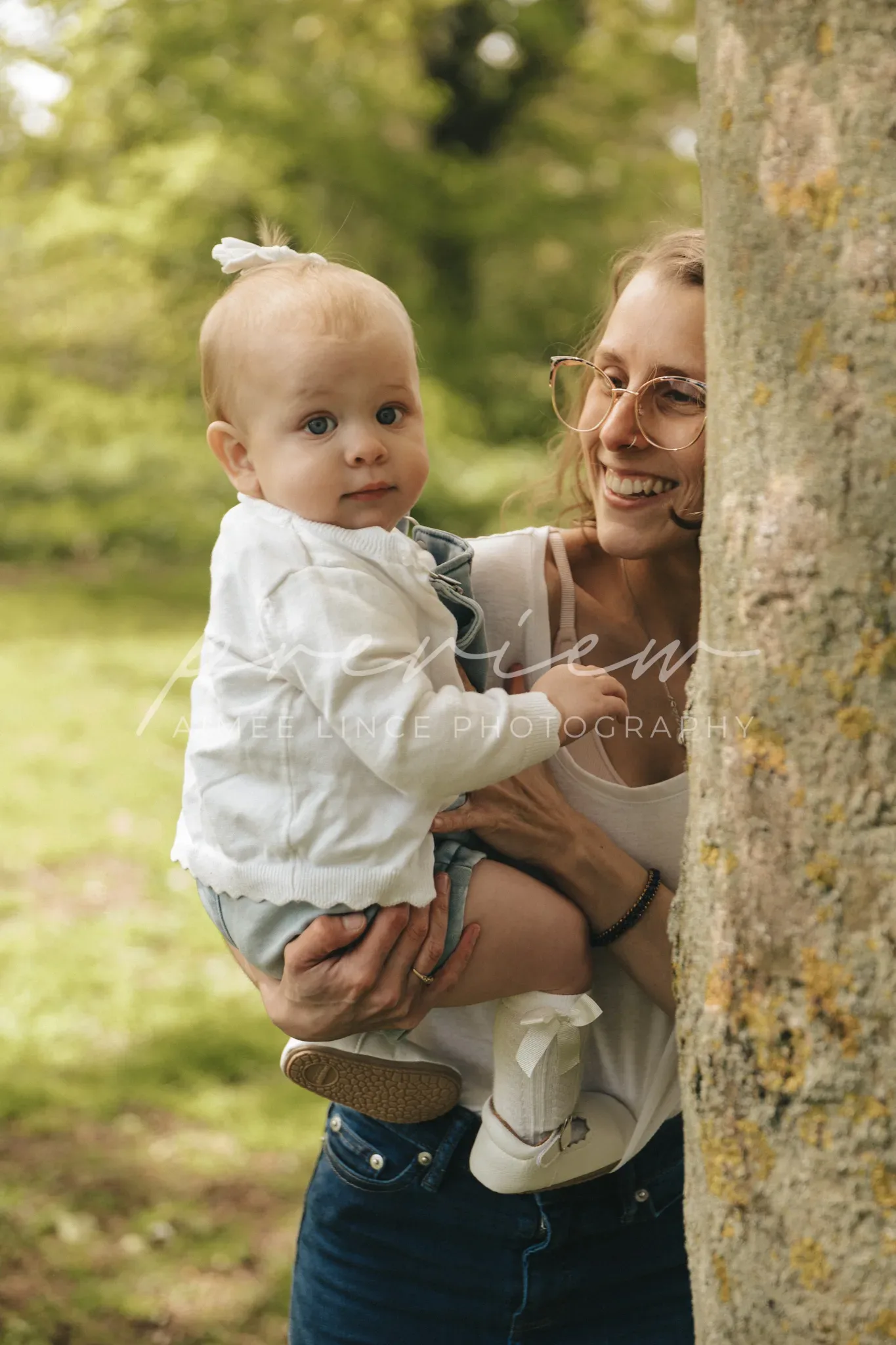 A joyful young woman with glasses and a ponytail, wearing a white top, holds a baby wearing a white shirt and denim. they are near a tree in a lush park, the baby looks at the camera while the woman smiles broadly.