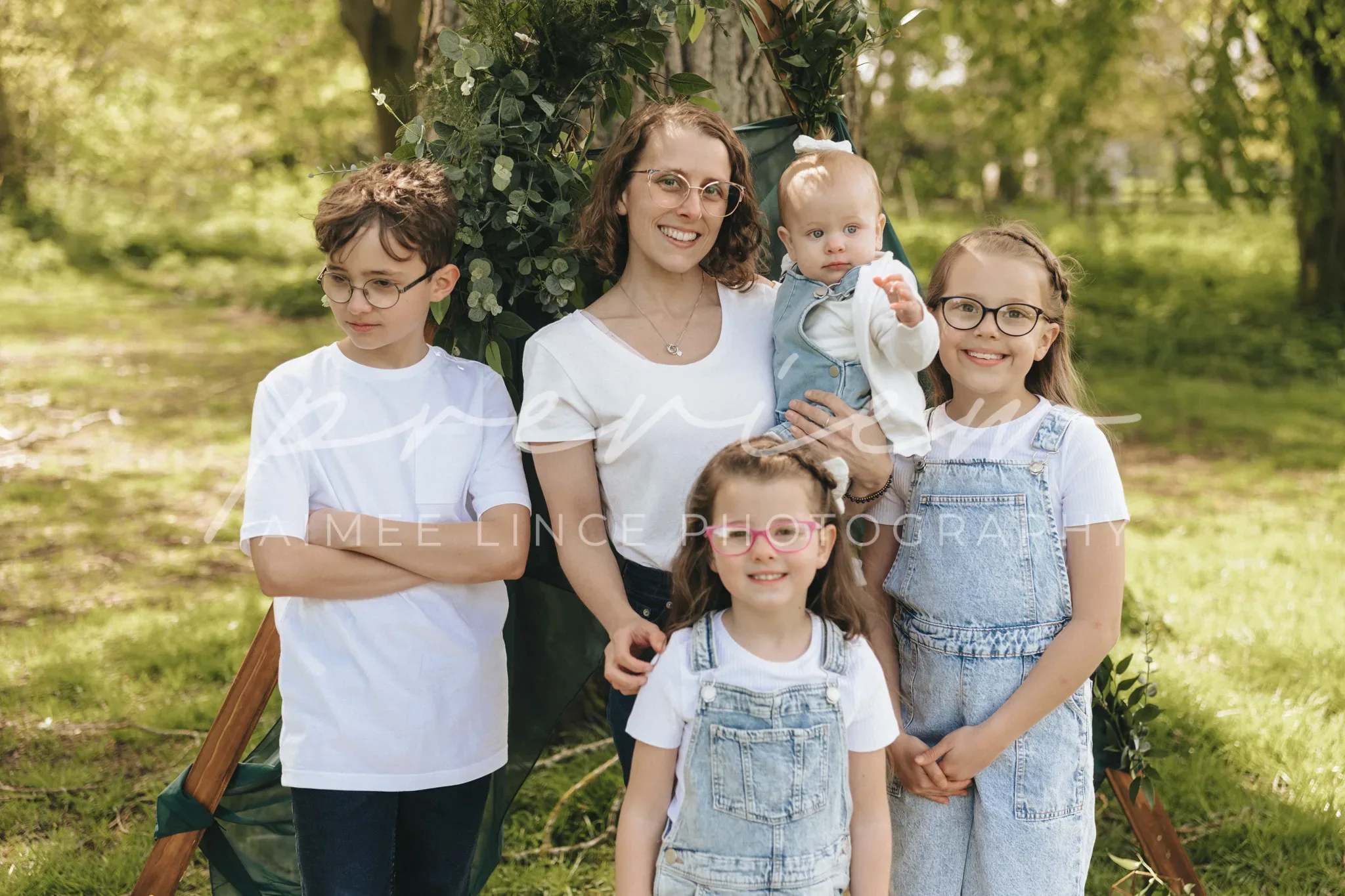 A happy family posing outdoors in a park. a mother holding a baby stands beside three children, two girls and a boy, all smiling. the setting features lush greenery and soft sunlight.