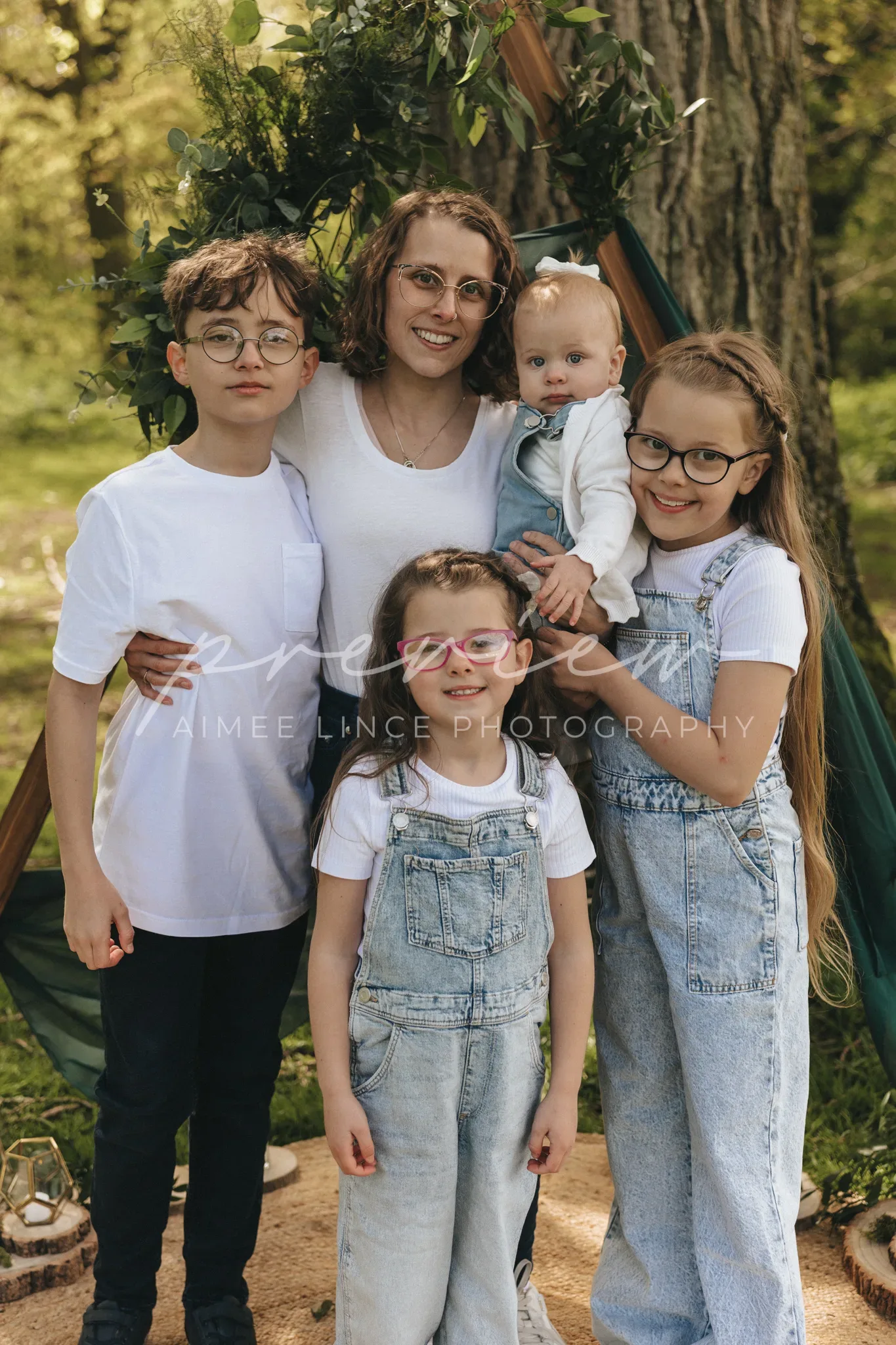 A family of five posing outdoors in a park, with two teenagers in glasses, a mother holding a baby, and a young girl, all wearing denim and white shirts, near a tent and trees.