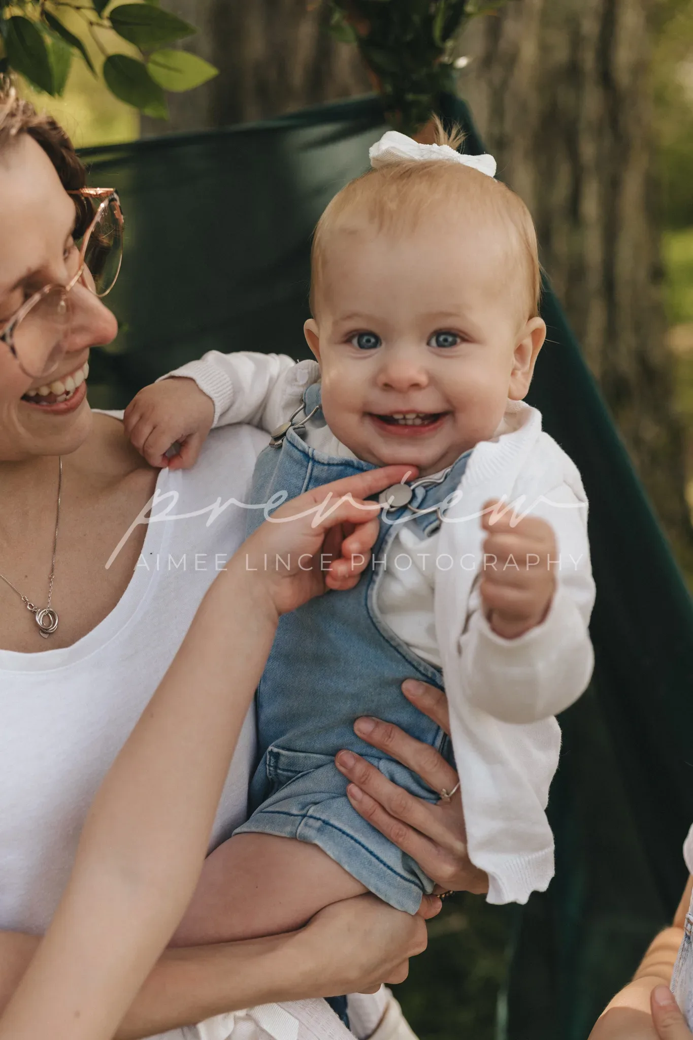 A joyful young woman with short brown hair smiles while holding a laughing baby dressed in a white shirt and denim overalls. they are outdoors, with trees in the background. the text "aimee lincé photography" is overlayed.