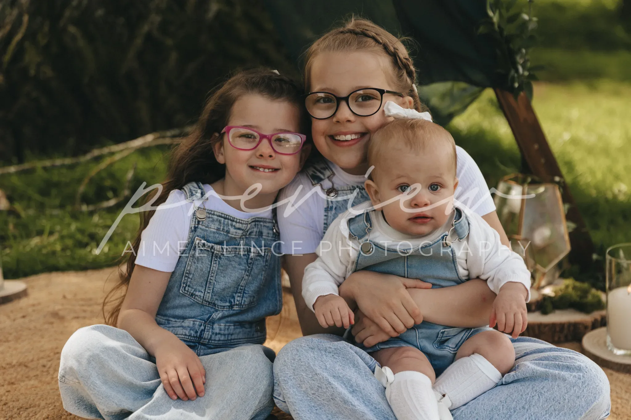 Two young girls and a baby sit outdoors. the older girls, wearing glasses and denim overalls, smile broadly. the baby, also in denim, is held by one of the girls. a picnic setup with a tent and a rug is visible behind them.