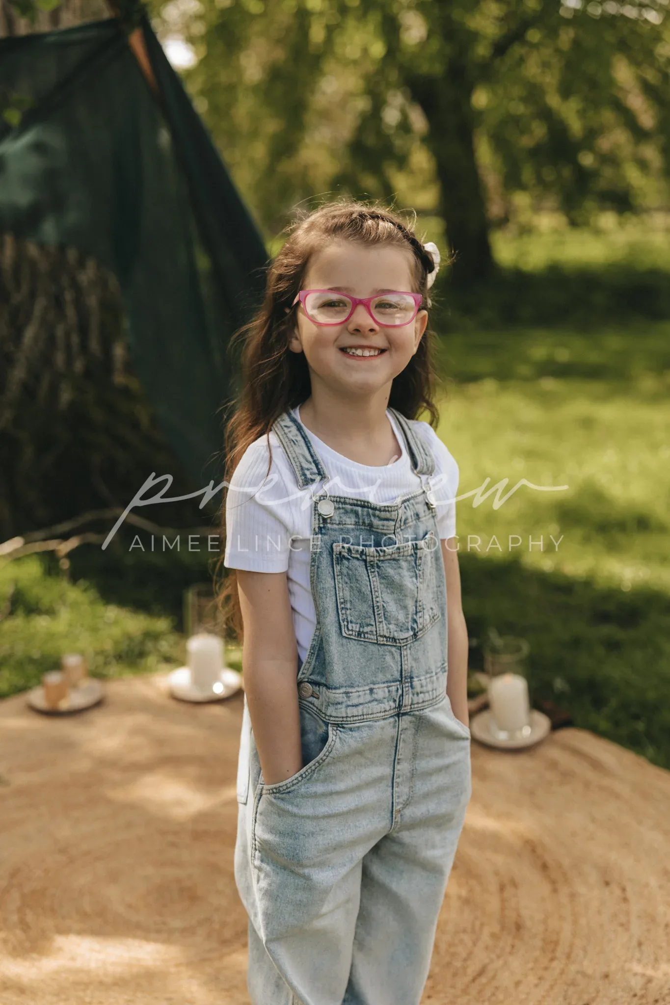 A joyful young girl with long brown hair and pink glasses wears denim overalls. she stands in a sunny park with a tent and picnic setup in the background, smiling broadly at the camera.