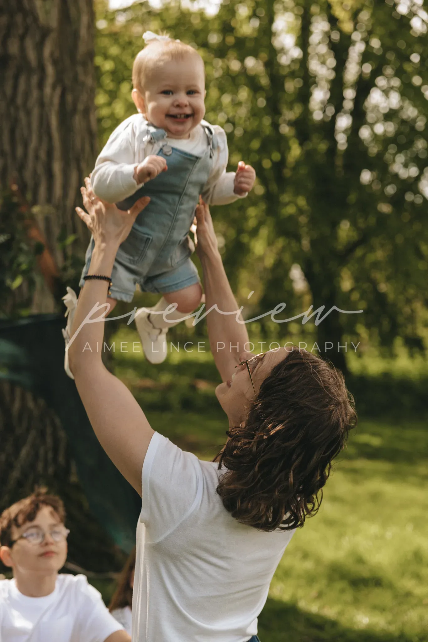 A joyful toddler in denim overalls is lifted high by a smiling woman in a white shirt, outdoors in a sunny park with trees. a young boy watches from the background.