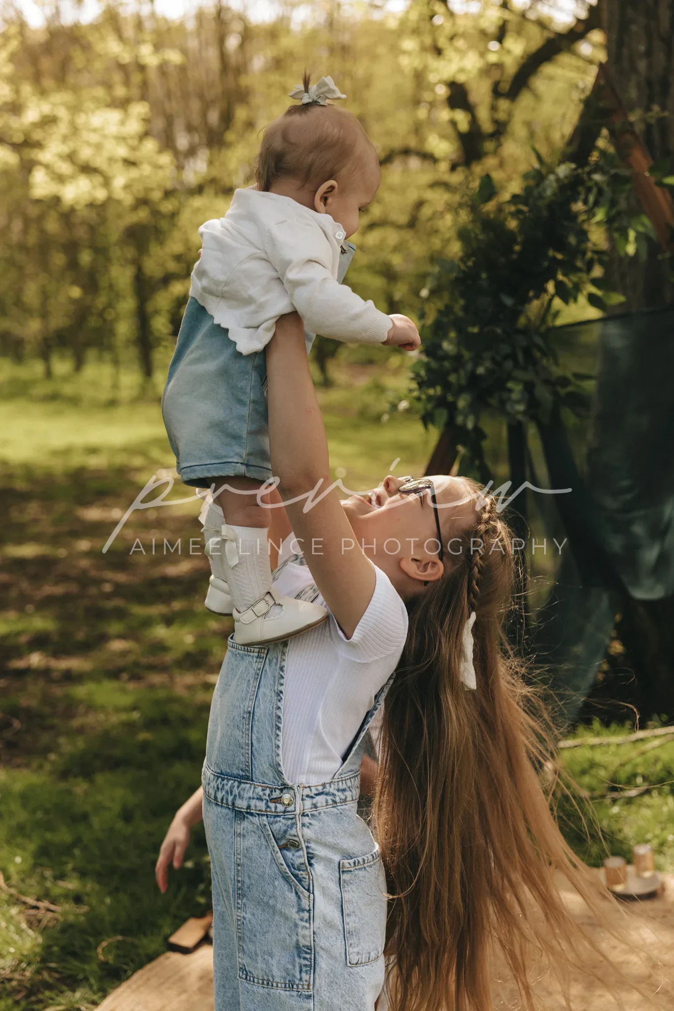 A woman in a denim overall lifts a baby girl into the air in a sunlit park. the child, wearing a light blue jacket and white leggings, smiles joyously. long shadows and lush greenery surround them.
