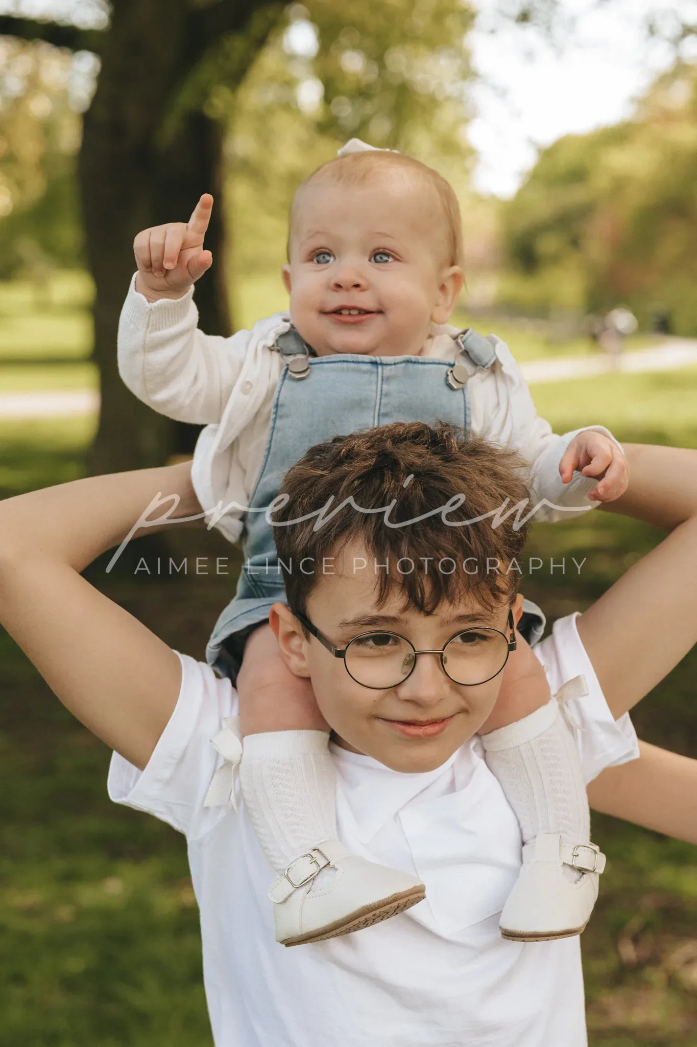 A joyful toddler sits on an older child's shoulders in a sunlit park. the toddler, dressed in overalls, points upwards, while the older child, wearing glasses and a white top, smiles broadly.