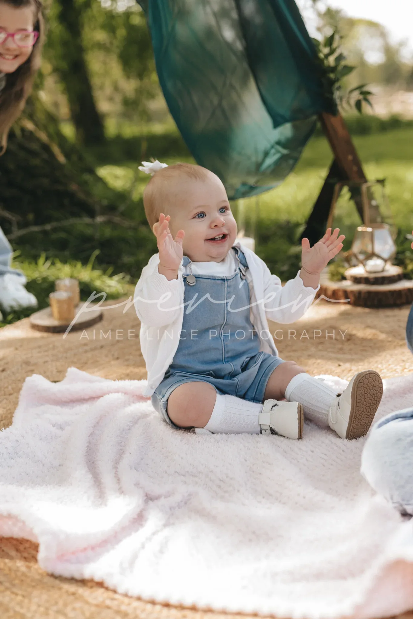 A joyful baby sits on a blanket, clapping hands, wearing a denim dungaree and white shoes. a smiling woman is partially visible in the background. soft, natural lighting enhances the serene outdoor setting with greenery.