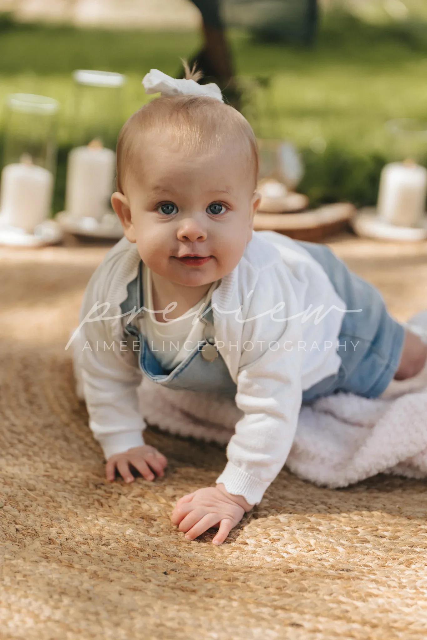 A baby with blue eyes and a white bow headband lies on a blanket, looking up with a joyful expression. they wear a denim shirt over a white top. soft sunlight filters through trees, creating a warm backdrop.