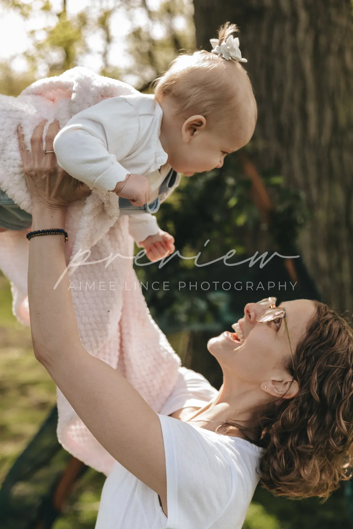 A joyful woman in a white shirt lifts a baby wearing a light pink outfit and a white bow headband above her head in a sunny, leafy park setting. the baby looks delighted and the woman is smiling broadly at the baby.