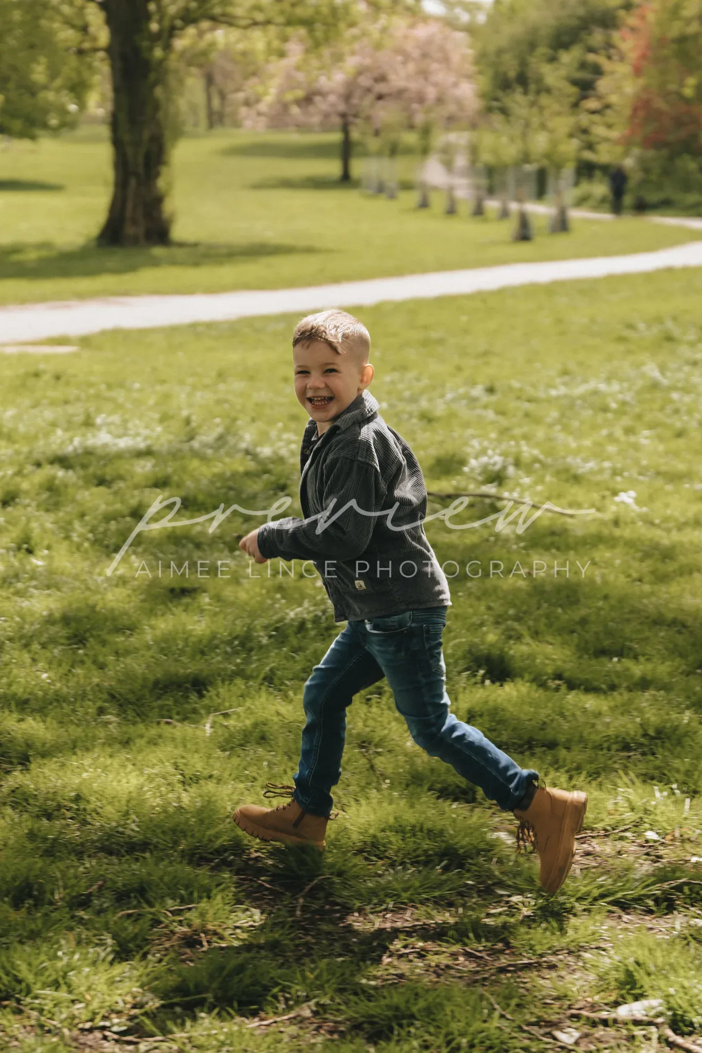 A young boy joyfully running on a grassy field in a park, with blooming trees and a path in the background. He's wearing a black jacket, blue jeans, and tan boots.