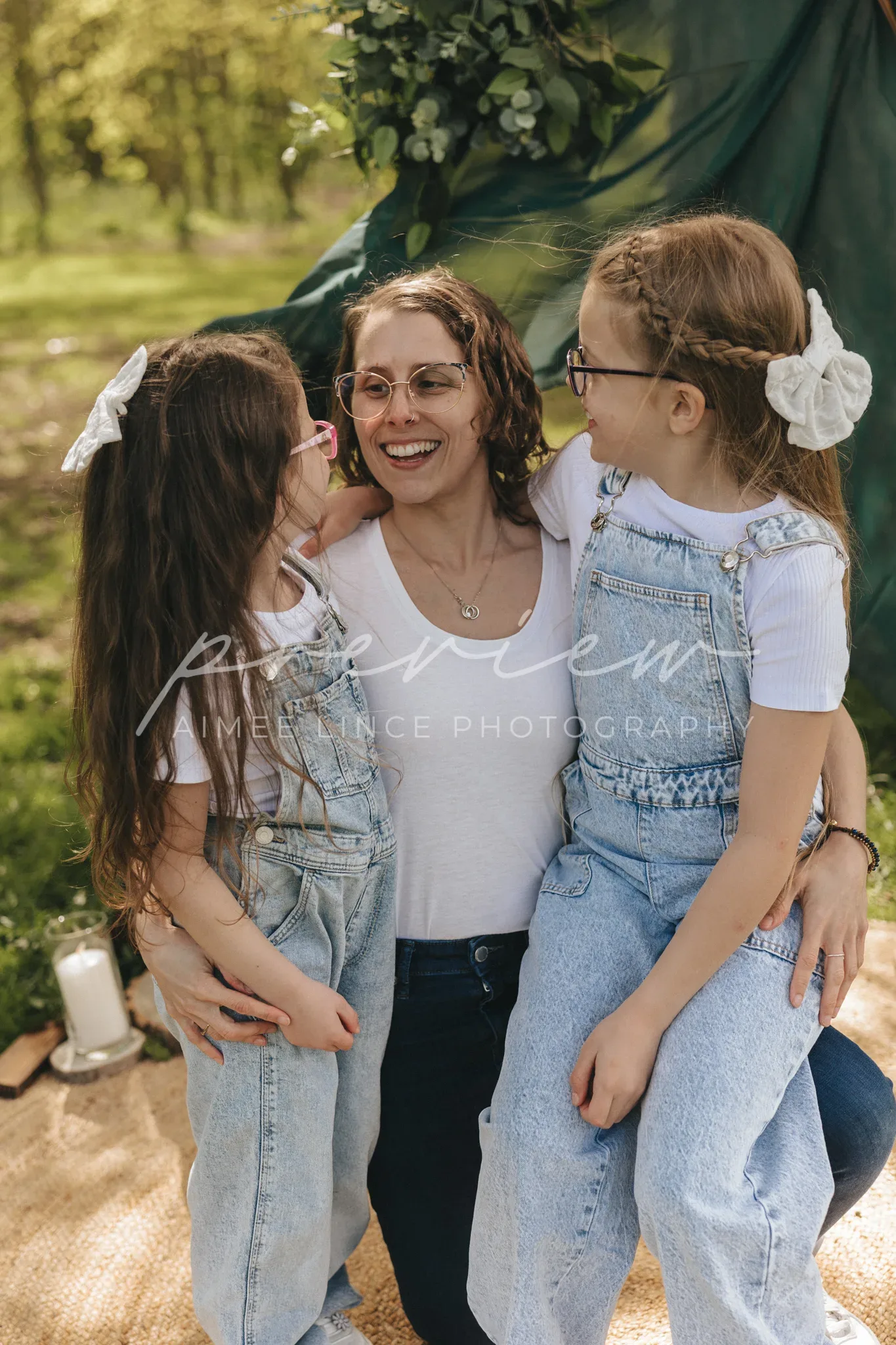 A joyful woman wearing glasses smiles, surrounded by two young girls in denim overalls. they are outdoors, near a tent under trees, with sunlight filtering through leaves. the woman embraces the girls as they share a happy moment.