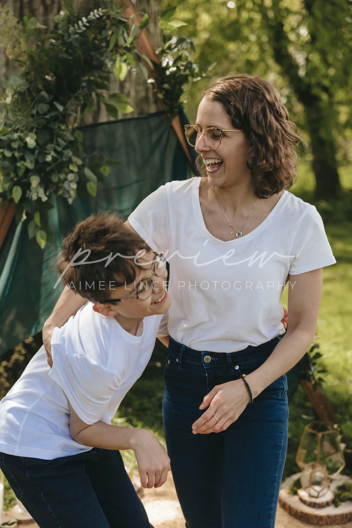 A woman and a young boy, both in white shirts and jeans, share a joyful moment in a sunny park. the boy leans against the woman, smiling broadly, as she looks down at him with a laugh. a tent and greenery are in the background.