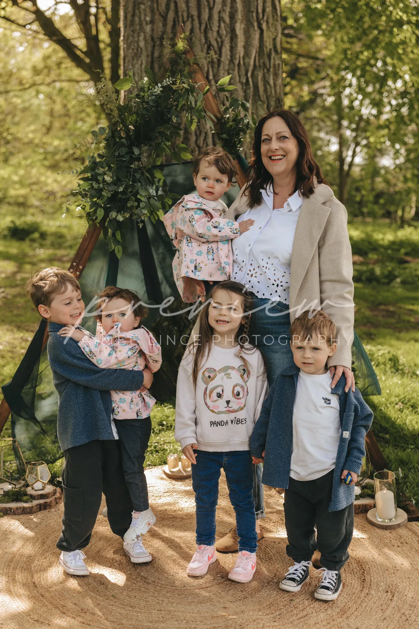 A joyful family portrait outdoors with a woman holding a baby, surrounded by four young children, three of which are peeking at the baby. they stand near a tree adorned with elegant greenery and flowers, under soft sunlight.