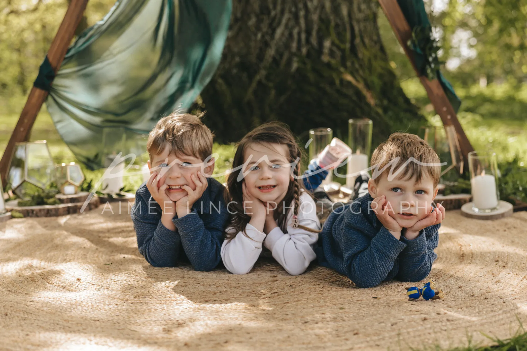 Three children lie on their stomachs under a hammock in a sunny park, with a picnic setup around them. they smile at the camera, resting their chins on their hands. the setting is serene and lush with greenery.