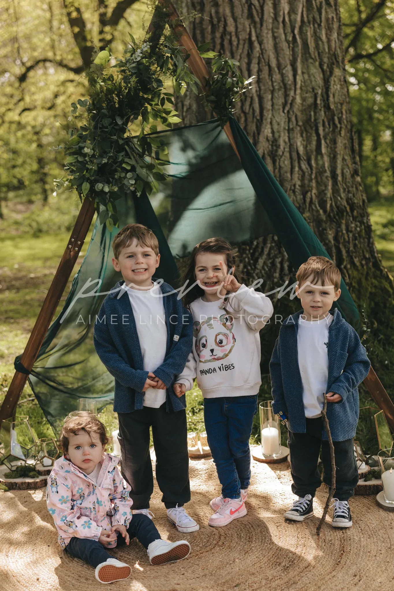 Four young children pose in front of a fabric teepee in a lush park. two boys, one making a peace sign, and a girl stand smiling, while a toddler girl sits in front. the teepee is decorated with greenery.