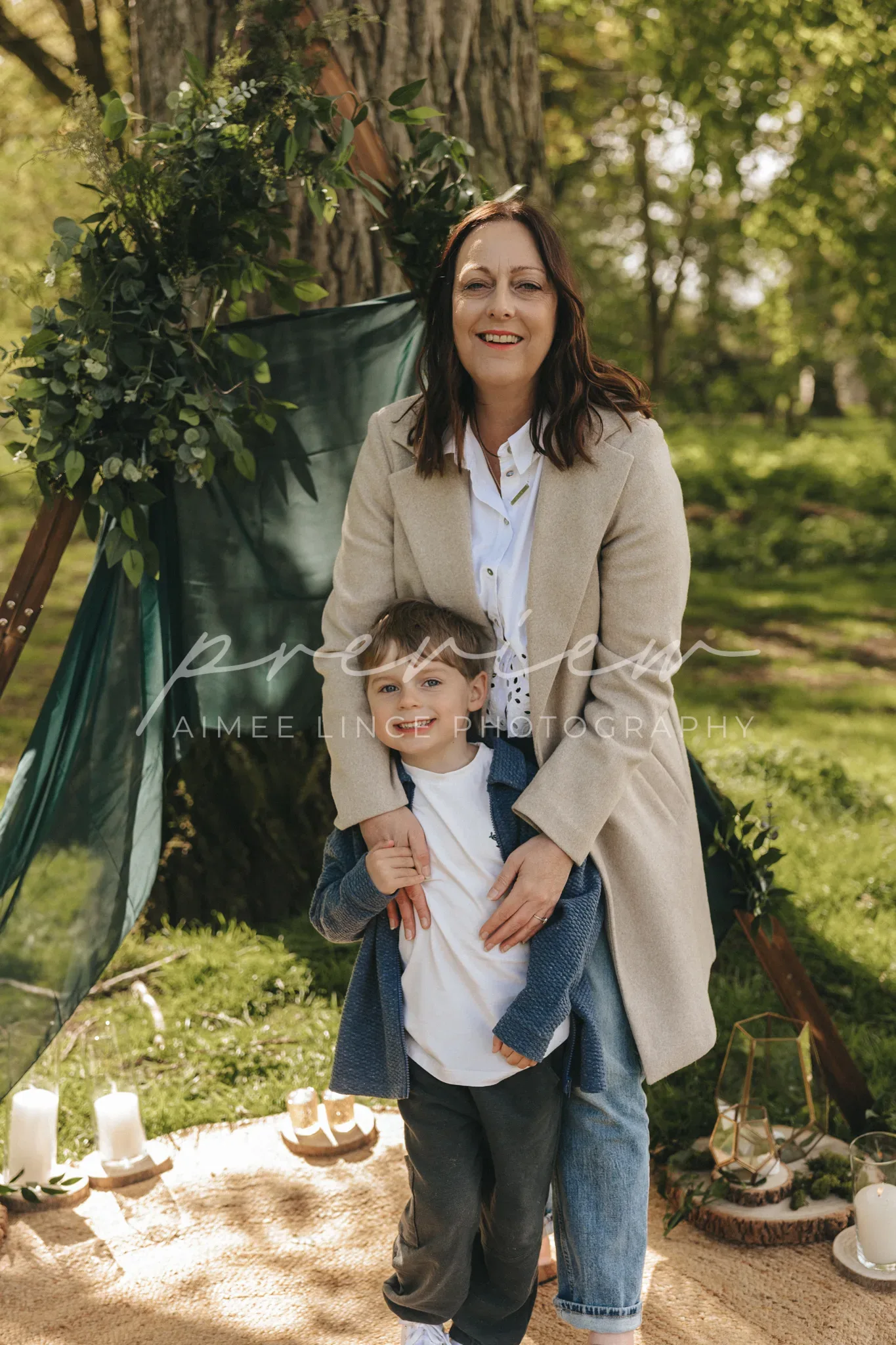 A woman and a young boy smile joyfully in a lush park, standing in front of a tree adorned with a whimsical green fabric. candles on the ground add a serene touch to the scene. both are dressed in stylish, casual outfits.