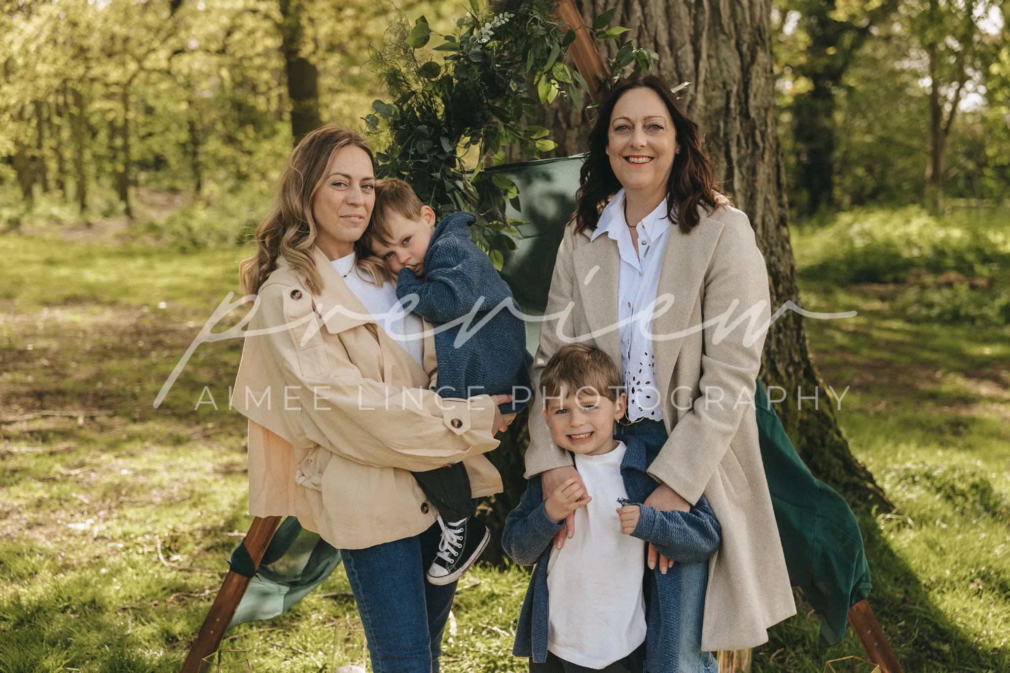Three generations of women and a young boy in a lush green park. the grandmother and mother stand smiling while one child hugs the mother and another stands in front. all are dressed in casual, comfortable clothing.