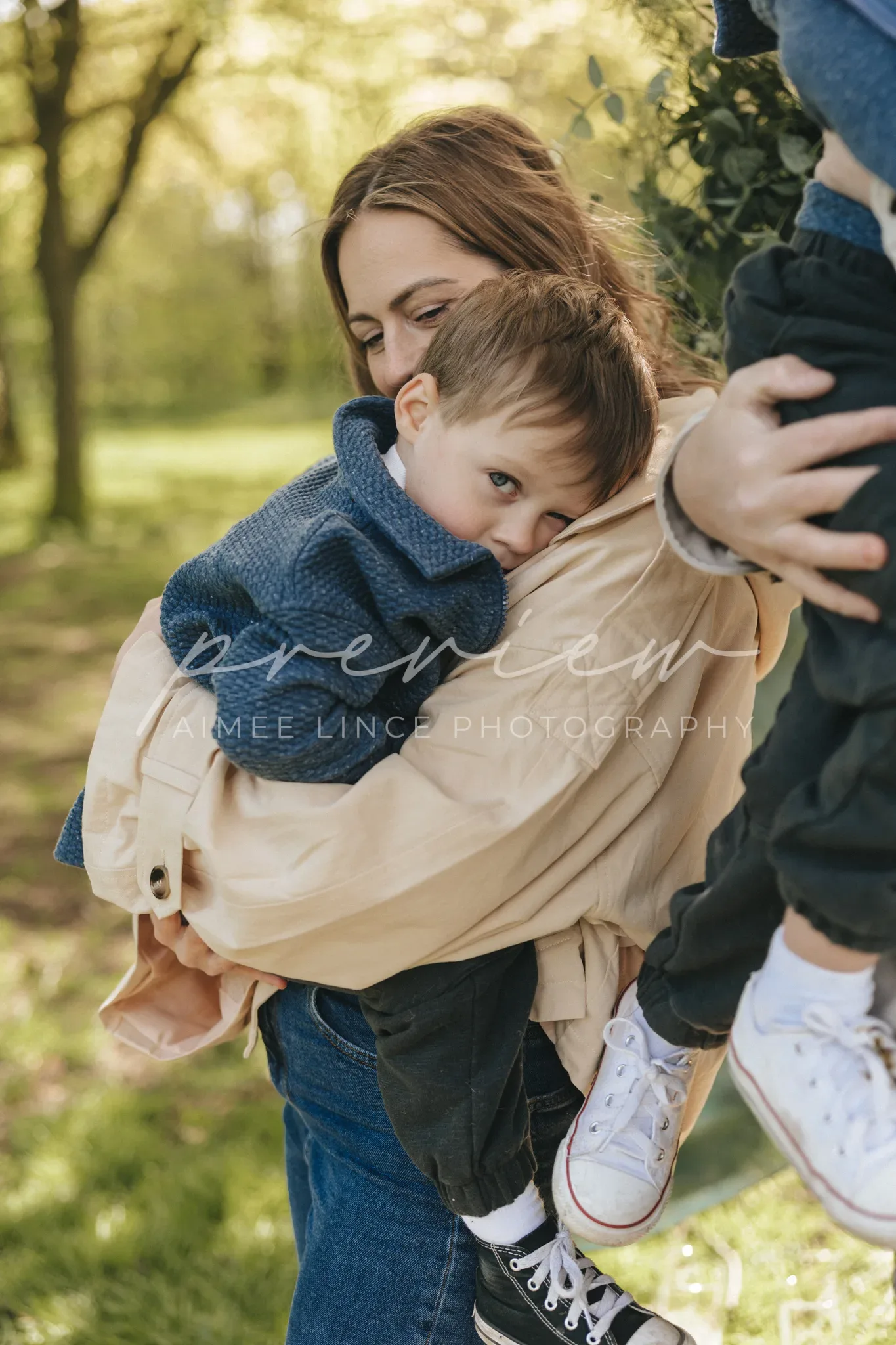 A mother lovingly holds her young son in a sunny park. the boy, dressed in a blue sweater and jeans, rests his head on his mother's shoulder while she, wearing a beige jacket, embraces him warmly. trees with green leaves fill the background.