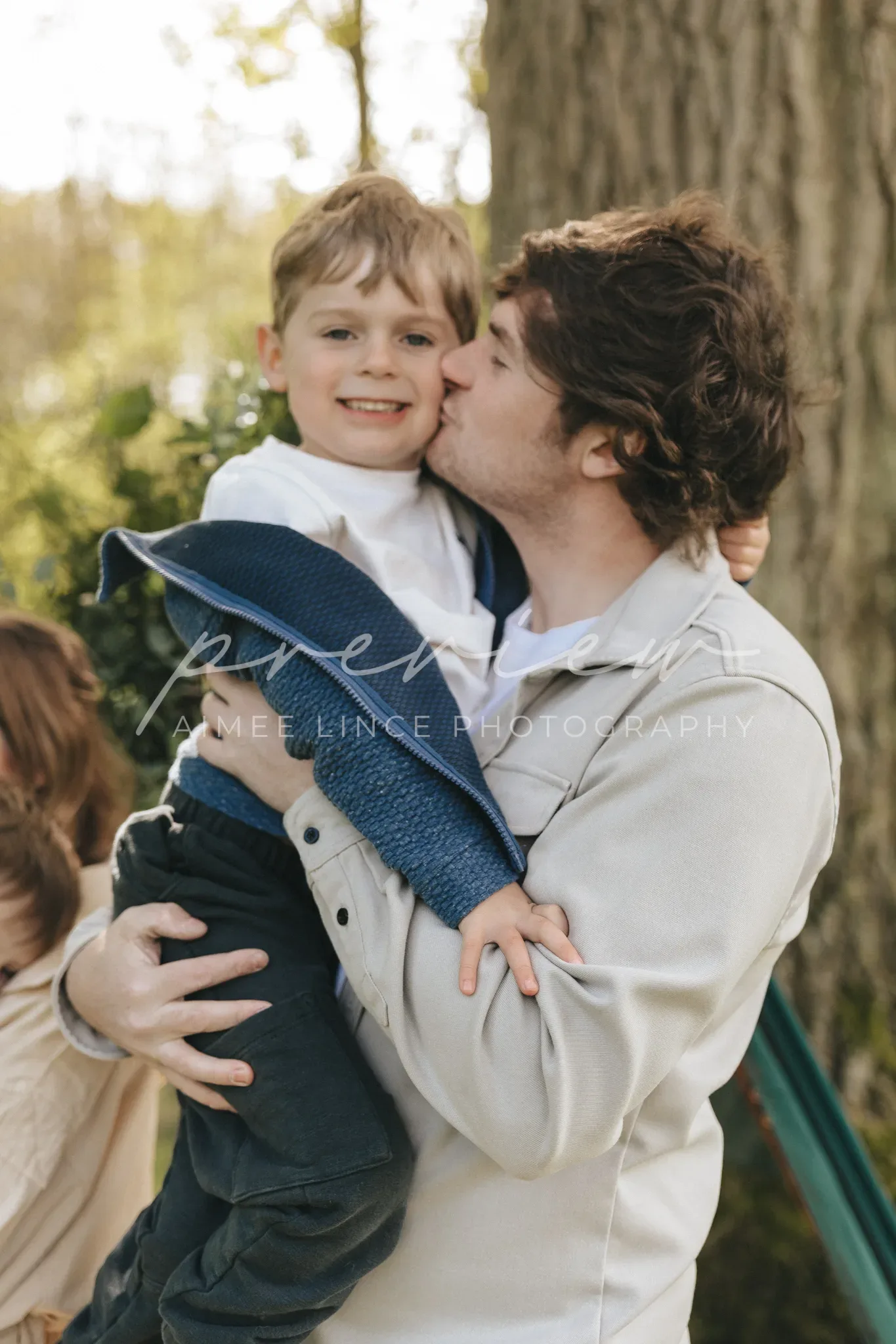 A father tenderly kisses his smiling young son on the cheek while holding him in his arms outdoors, with trees in the background illuminated by soft sunlight.
