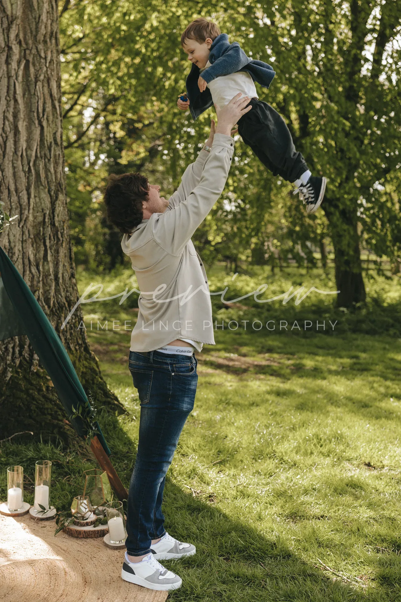 A woman, wearing a hoodie and jeans, playfully lifts a young boy in the air under a tree in a lush park. nearby, candles in glass jars are set on the ground beside a draped fabric. the setting is sunlit and verdant.