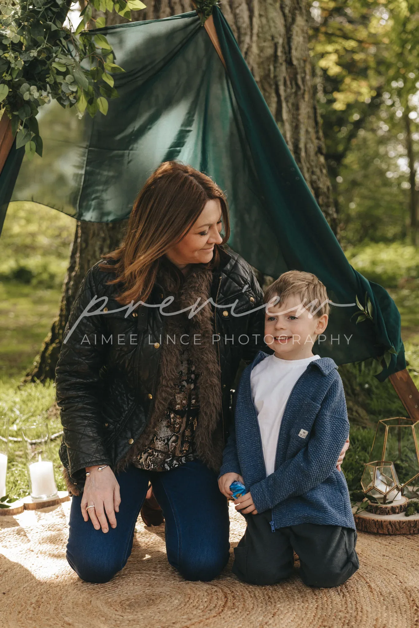A woman and a young boy kneel together under a makeshift tent in a forest. the woman, wearing a black jacket and blue jeans, lovingly looks at the boy who is dressed in a blue sweater and white shirt. soft sunlight filters through the trees around them.