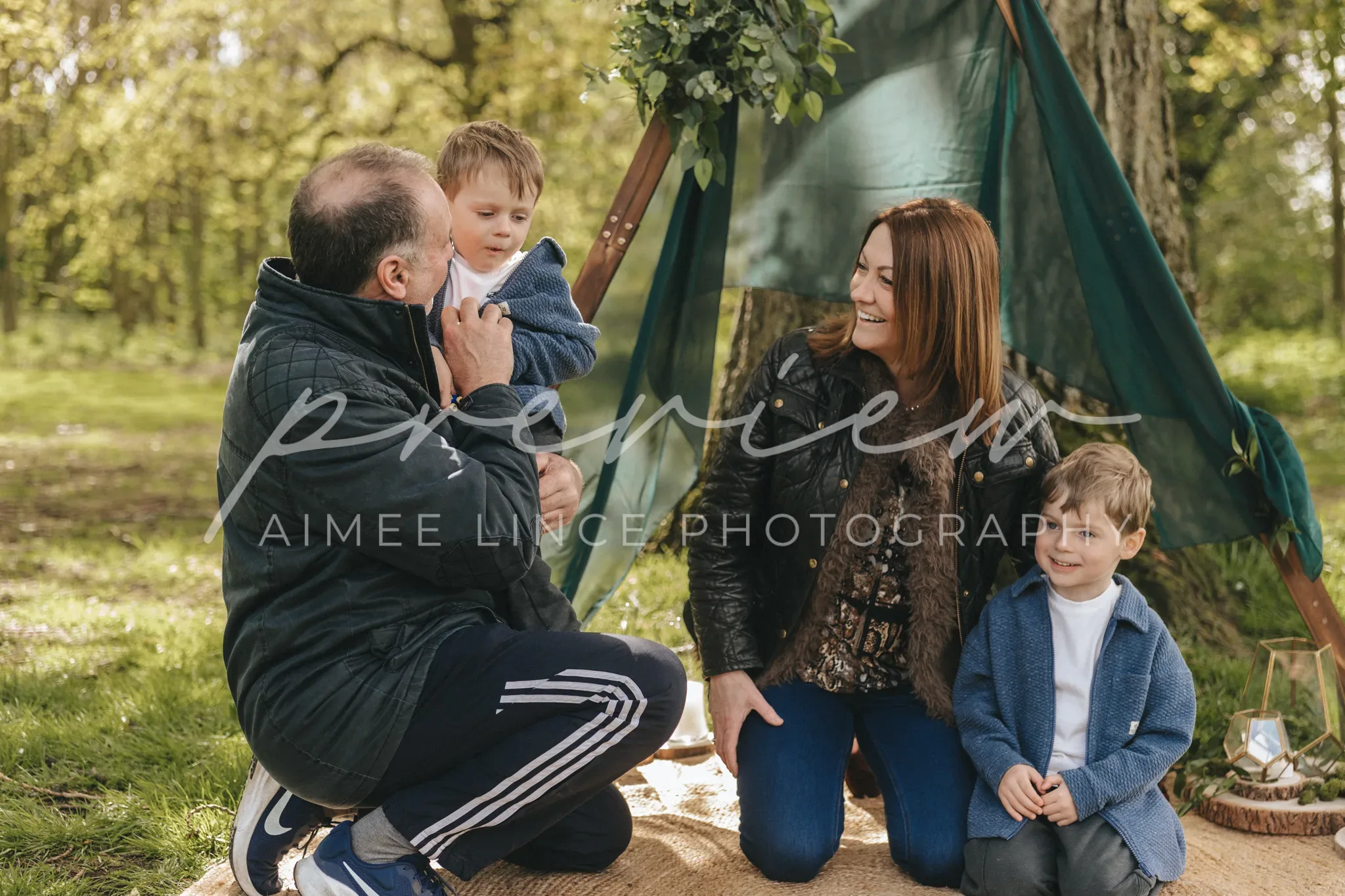 A family outdoors, with a father lifting a toddler, mother looking on smiling, and another young boy sitting beside them under a green teepee, in a sunlit wooded area.