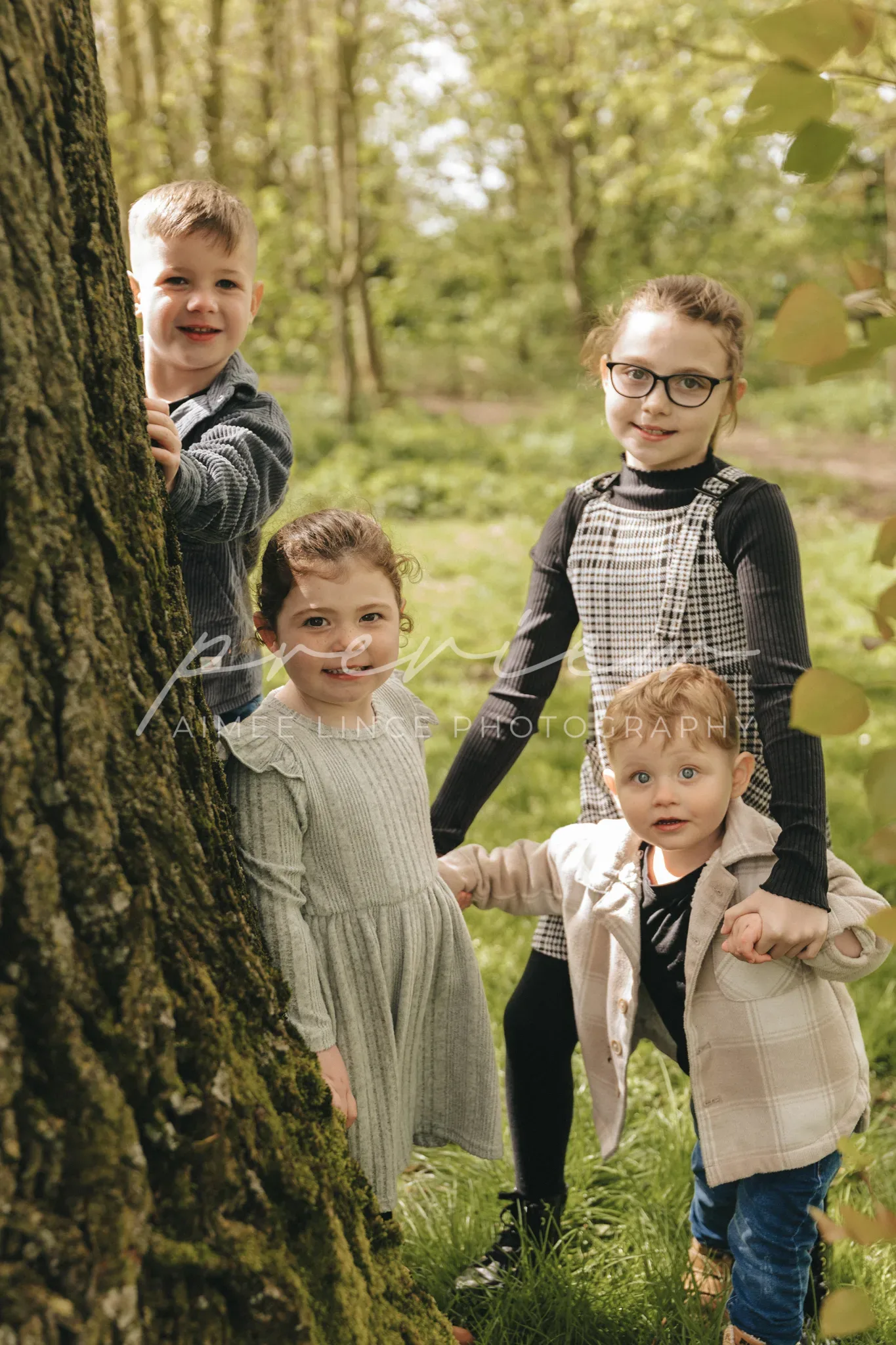 Four children of varying ages stand playfully behind a tree in a sunlit forest. The two girls, including Gabrielle, wear dresses and the two boys are in shirts and vests. They all smile