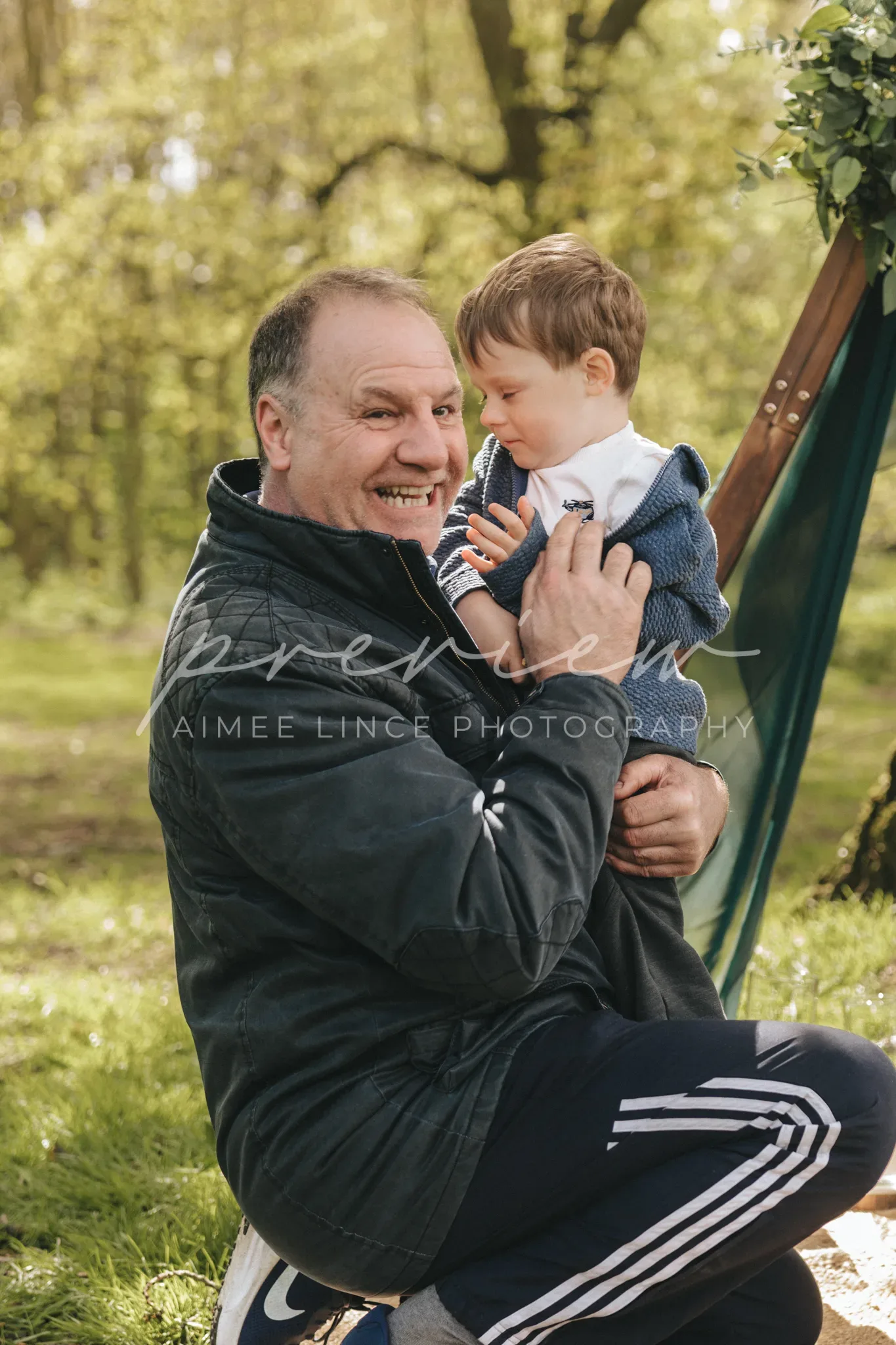 A joyful moment between a middle-aged man and a young child as they interact closely at a park. the man is crouching, holding the child in his arms, both are smiling, surrounded by lush green trees.