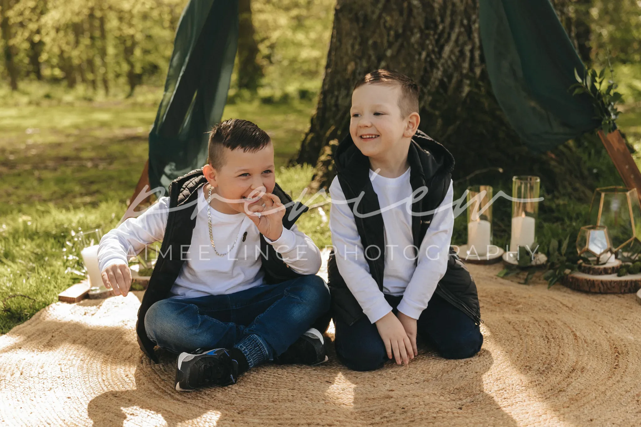 Two young boys sitting under an outdoor tent with a picnic setup, smiling and enjoying a sunny day. one boy eats a marshmallow. they wear white shirts, vests, and jeans in a grassy park.