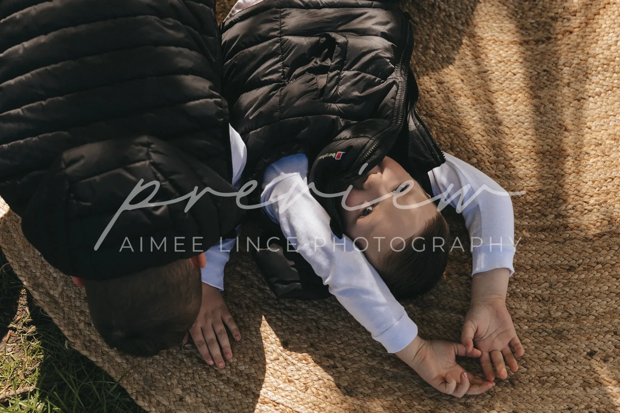 A child lies on a woven blanket outdoors, partly covered by a black puffer jacket. the sun casts shadows of nearby leaves across the scene, adding a serene and natural ambiance.