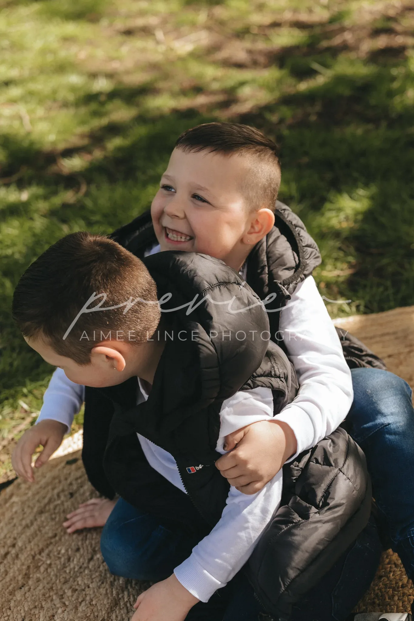 Two young boys laughing joyfully while sitting close together on a log outdoors. both wear dark padded vests over light long-sleeved shirts. the background shows sunlit grass. there's a watermark "aimee vince photography" on the image.