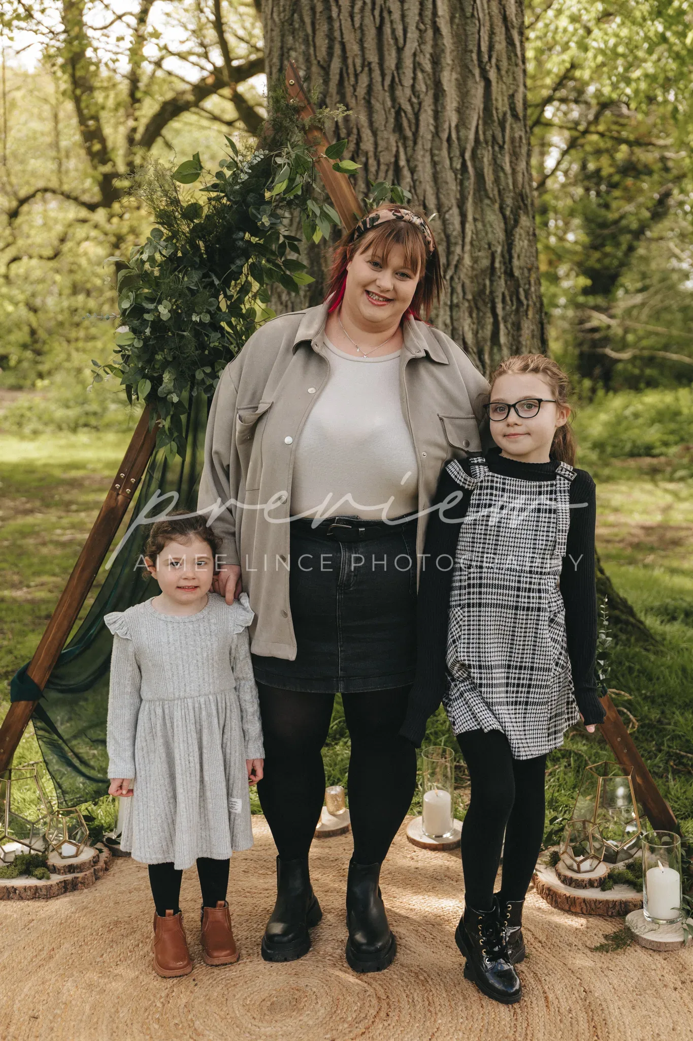 Gabrielle stands between two young girls in front of a large tree, smiling at the camera. A wooden tripod with greenery is on the left, and lanterns are on the ground. The