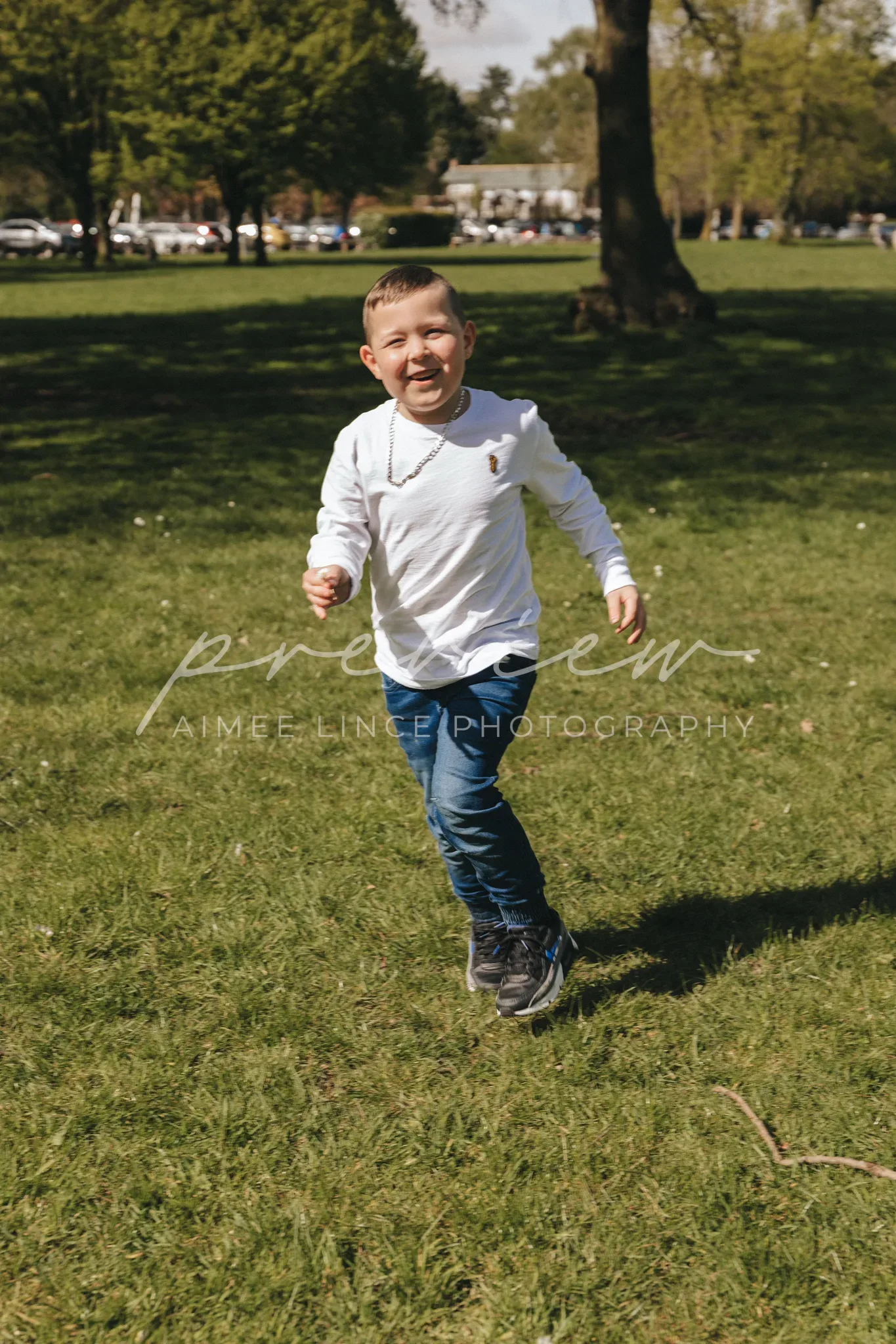 A young boy joyfully running towards the camera in a sunny park. he's wearing a white long-sleeve shirt, jeans, and sneakers, with a blurred background of green grass and trees. the image bears a watermark "aimee linen photography.